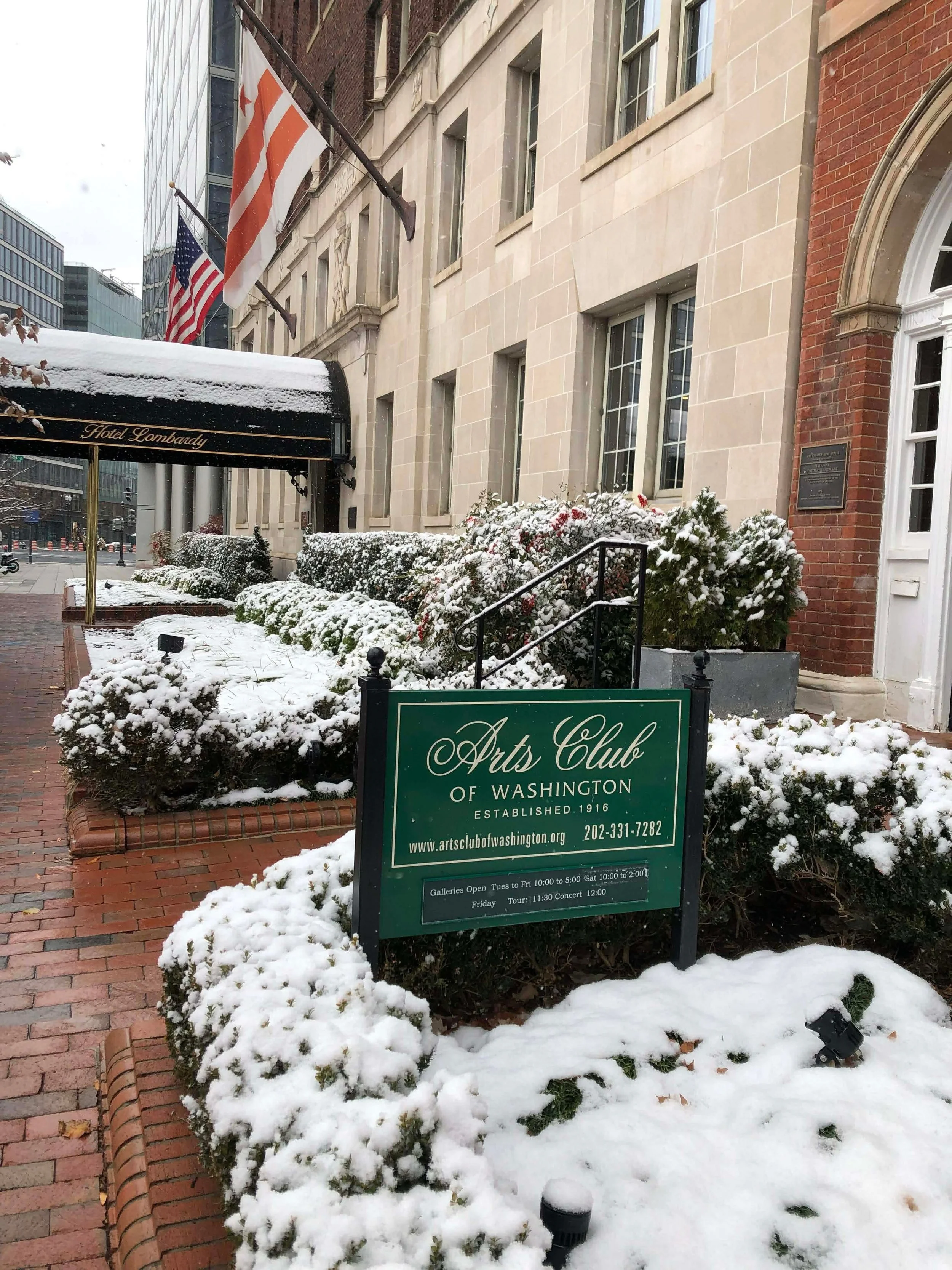 Snow-covered bushes and brick sidewalk in front of the Arts Club of Washington building, with American flags and building signs visible.