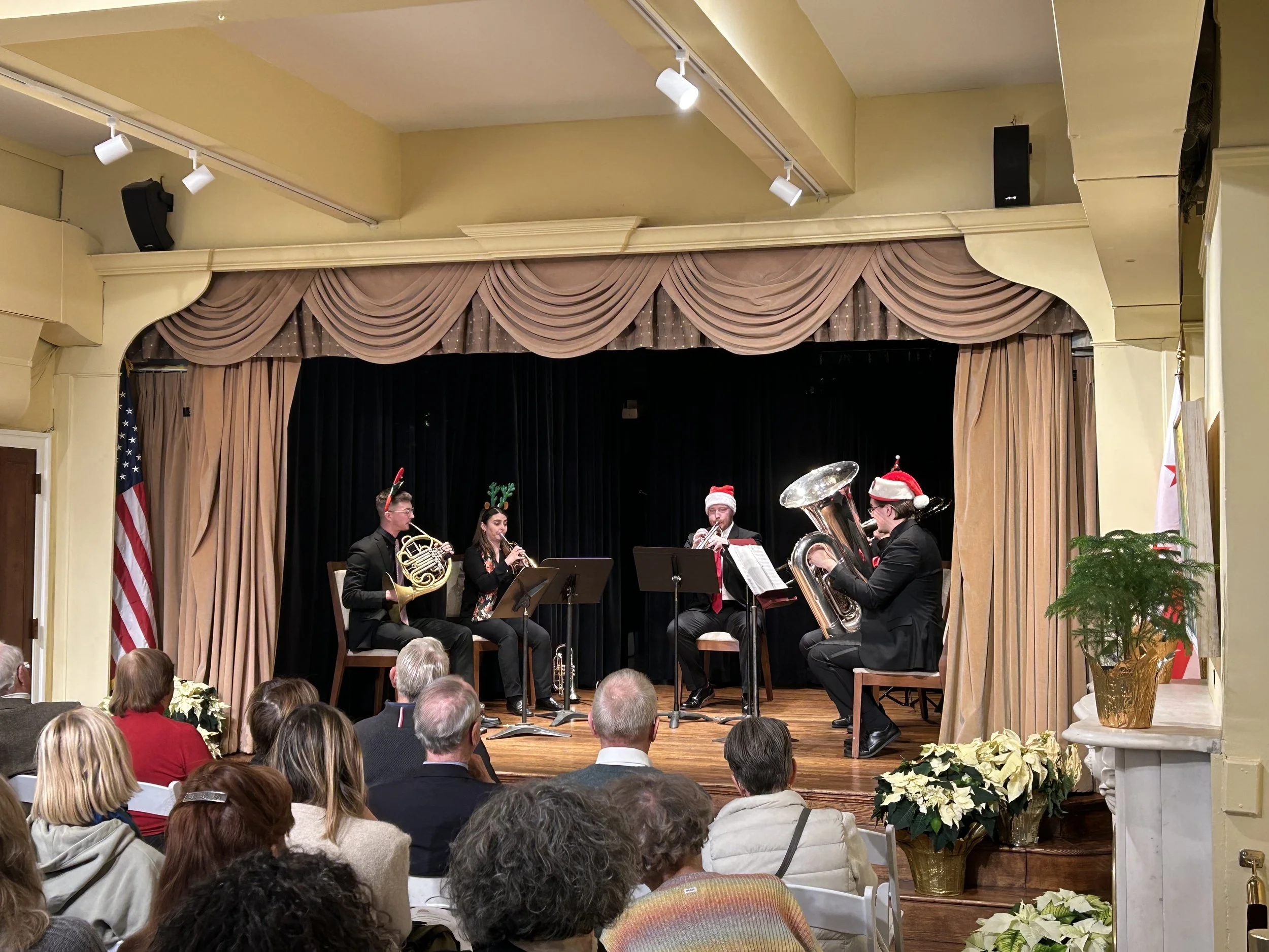 A Christmas-themed musical performance on a small stage with four musicians playing wind instruments. All are dressed in black and wearing holiday accessories such as Santa hats and reindeer antlers. The audience is seated in front of the stage, and the room is decorated with poinsettia plants and holiday decor.