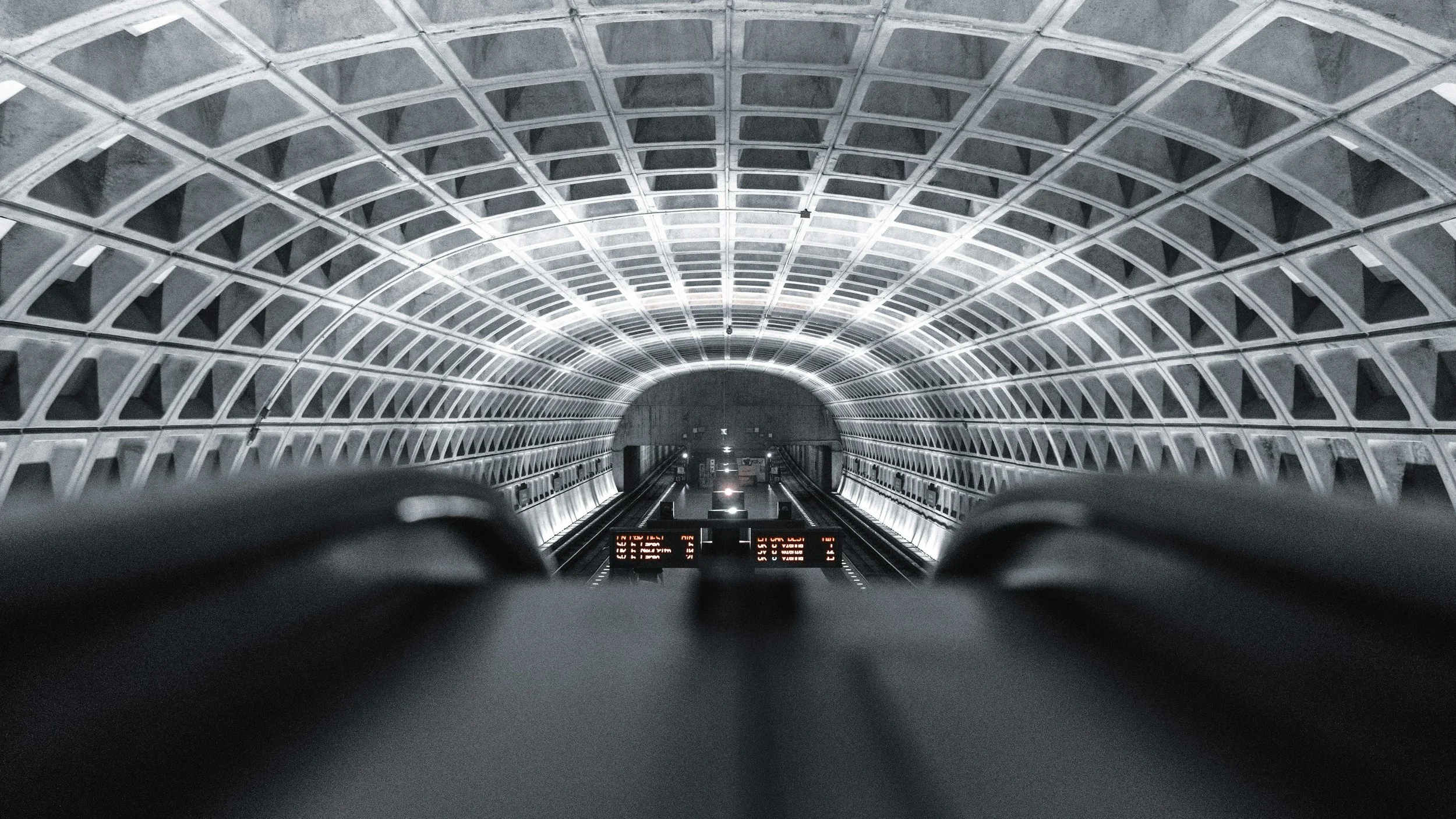View inside an underground subway tunnel, looking down the tracks towards the tunnel's arched ceiling with illuminated white lines.