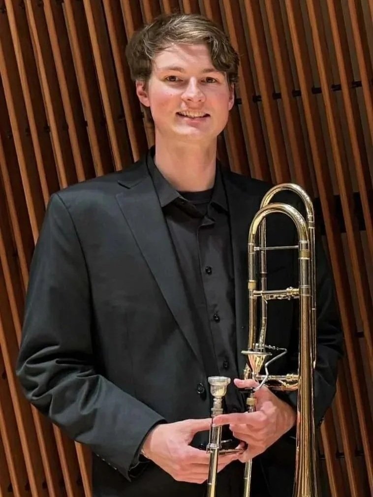 Young man in black suit holding a trombone standing in front of a wooden slat wall.