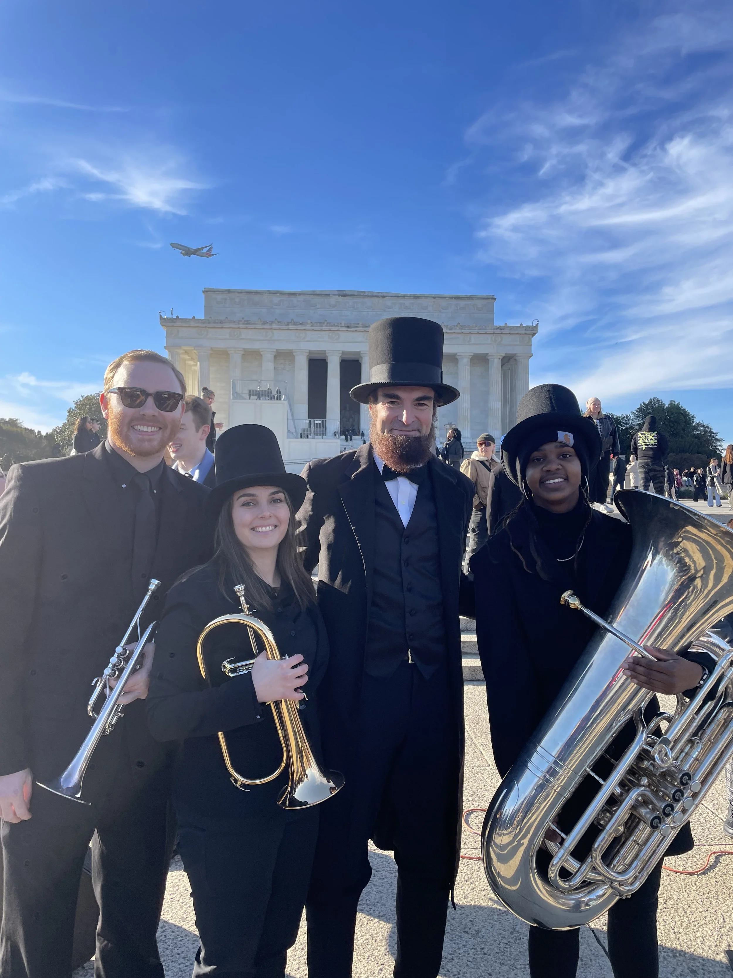 Group of four musicians dressed in black, standing in front of the Lincoln Memorial with an airplane flying overhead in clear blue sky.