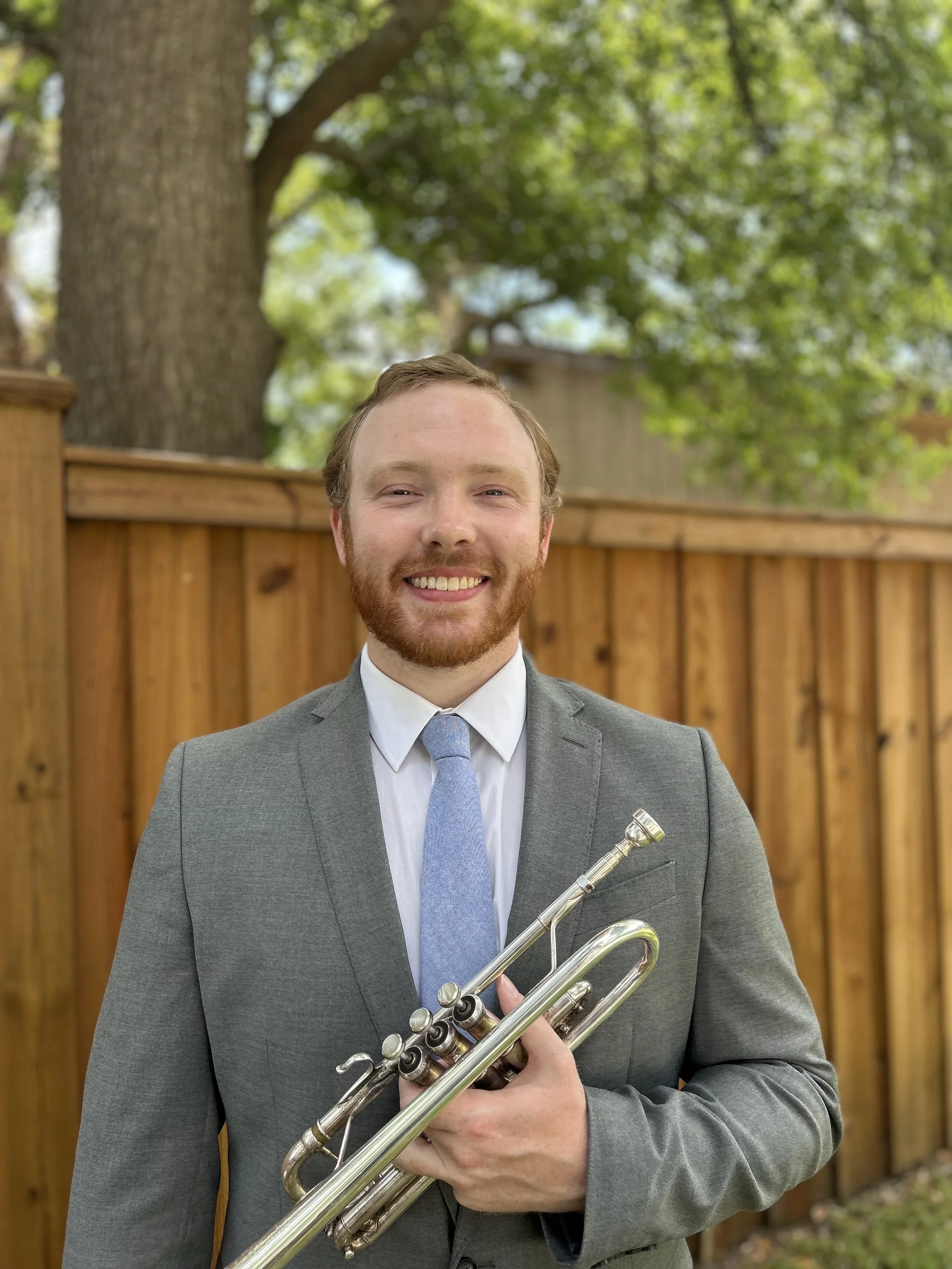 A man with a beard and a wedding ring, dressed in a gray suit and white shirt, smiling while holding a brass trumpet outdoors.