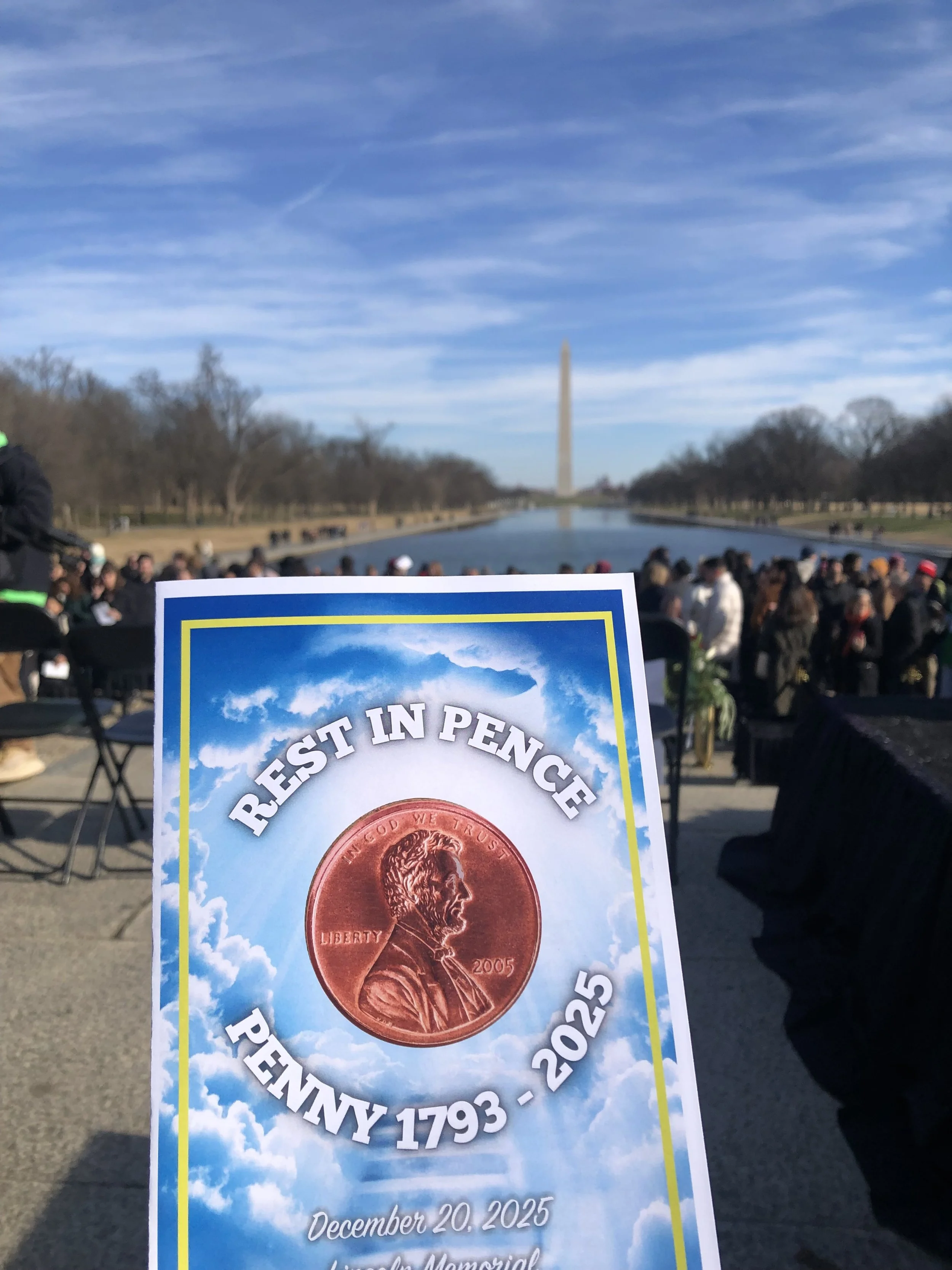 A pamphlet titled 'Rest in Pence Penny 1793-2025' with a penny image on it, held in front of the Lincoln Memorial Reflecting Pool with the Washington Monument in the background, and a large crowd gathered around.