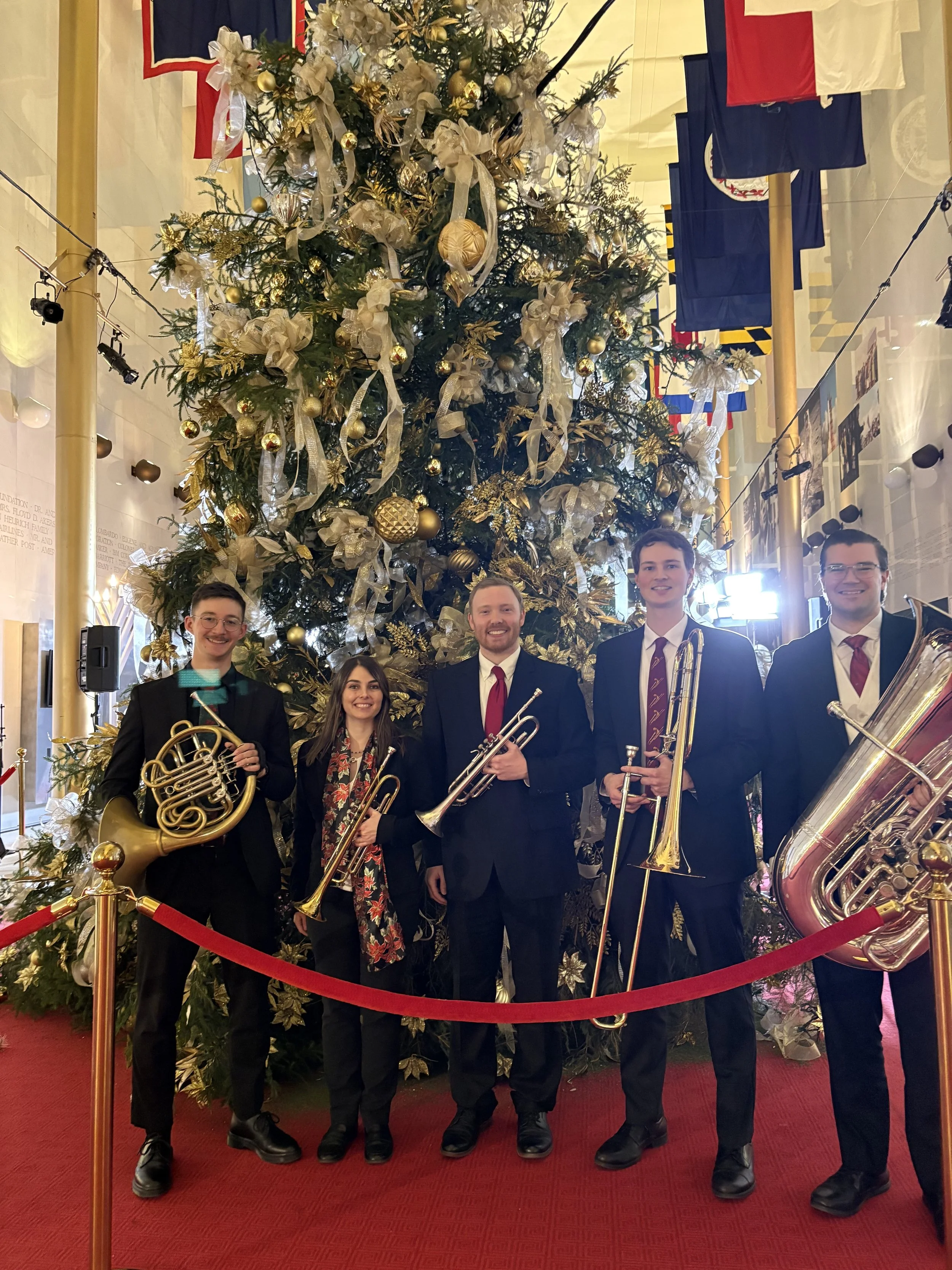 Six people dressed in formal attire holding musical instruments standing in front of a large decorated Christmas tree at a holiday event.