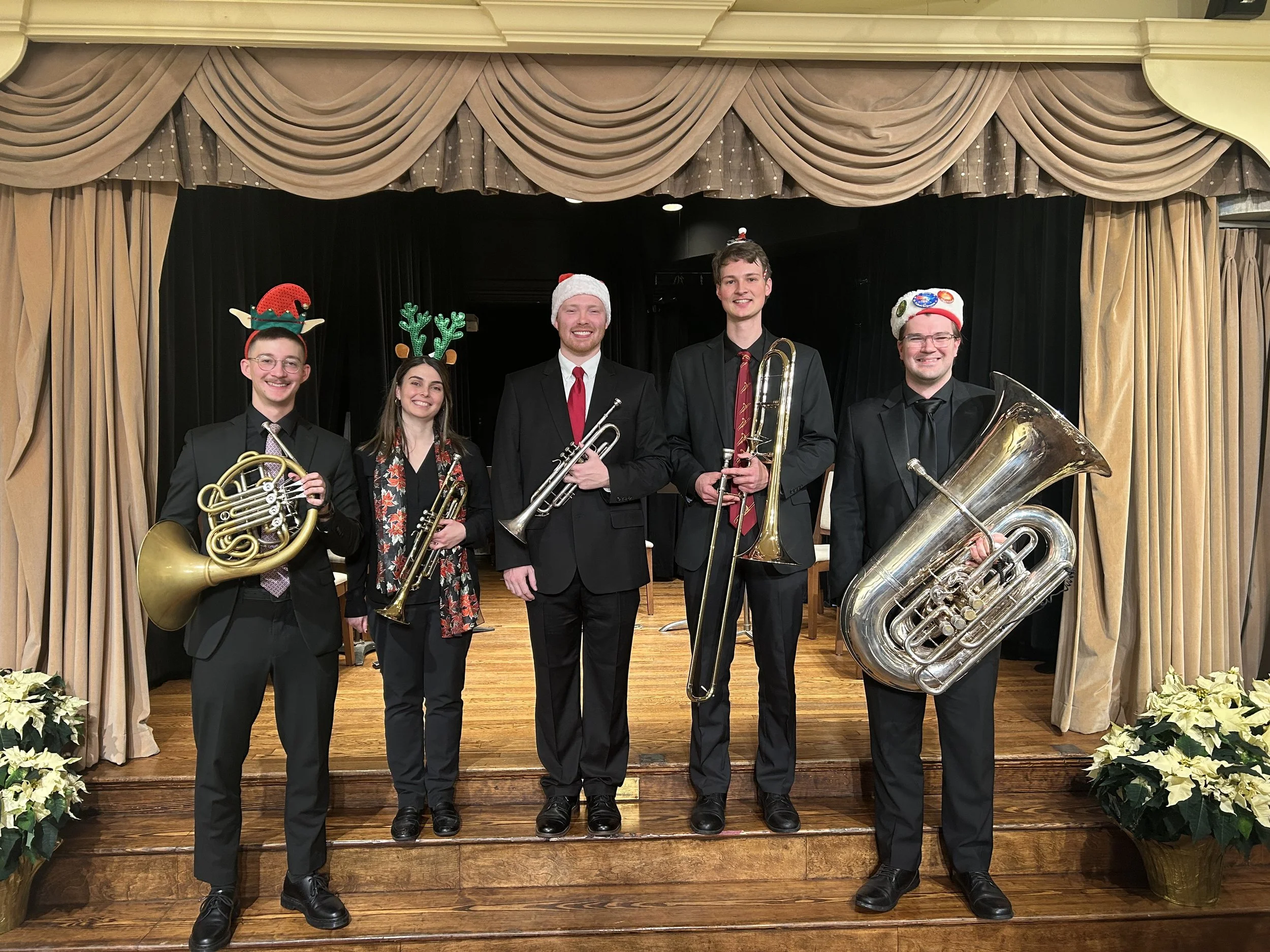Group of five musicians dressed in formal black suits, standing on stage with Christmas-themed hats and reindeer antlers, holding musical instruments including French horn, trumpet, trombone, and tuba, in a decorated performance venue with curtains and poinsettia plants.