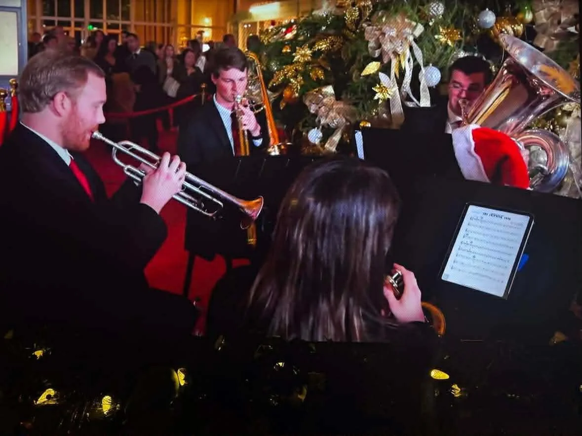 A group of musicians playing brass instruments at a Christmas event, with a decorated Christmas tree in the background and an audience watching.