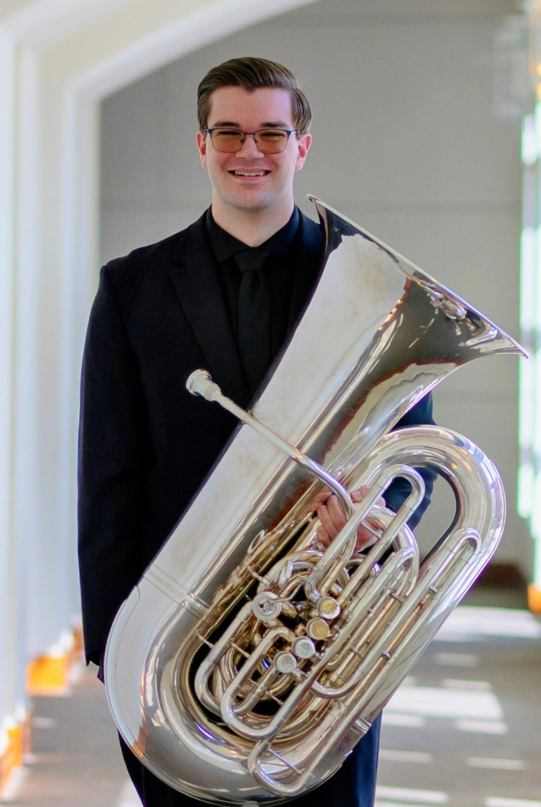A young man in a black suit and glasses smiling while holding a shiny brass tuba.