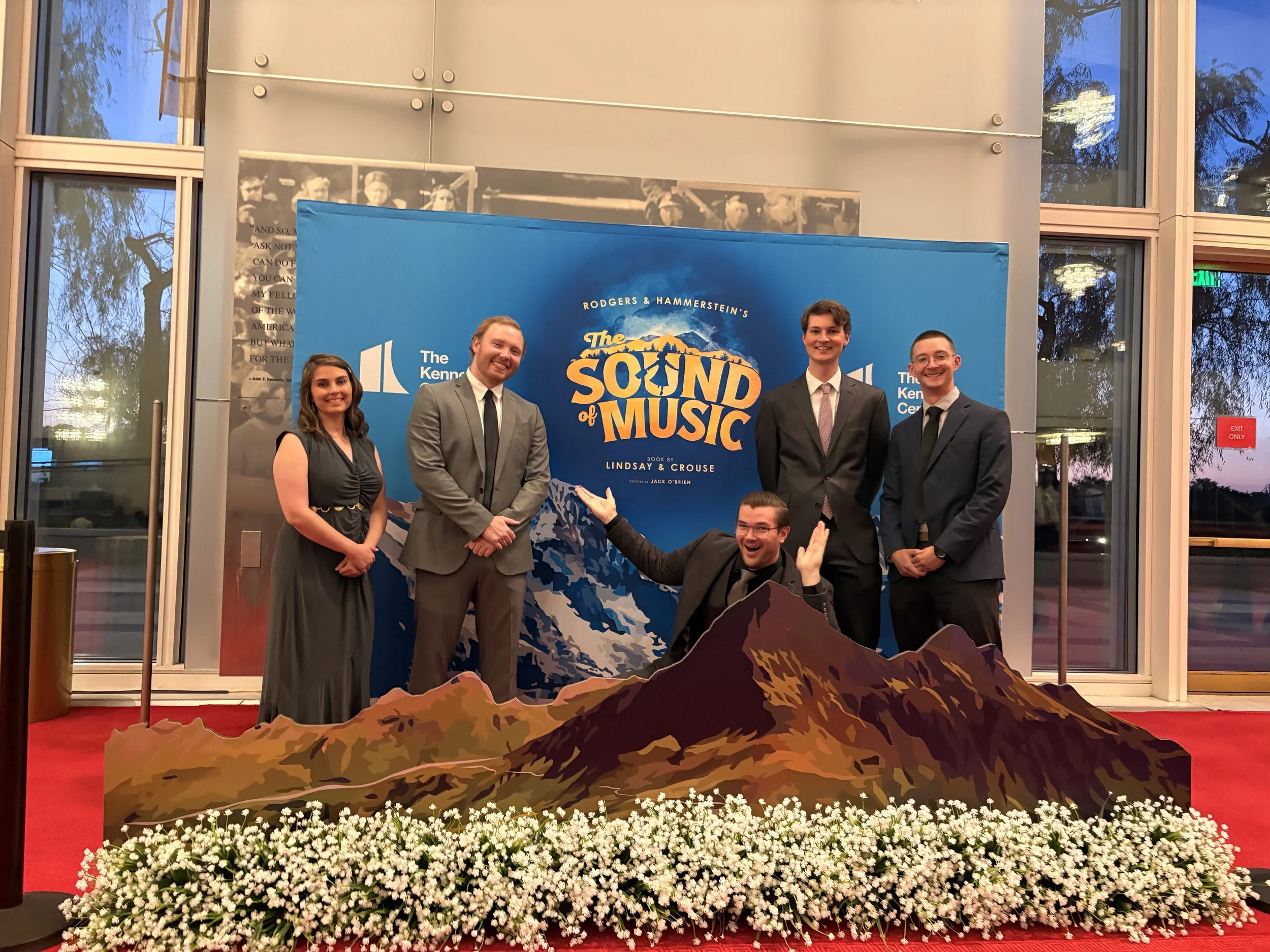 Six people are posing on a red carpet in front of a backdrop for the musical 'The Sound of Music'. They are dressed in formal attire, with four men and one woman standing and one man kneeling in front with arms raised. There is a mountain and flowers decoration in front of them.