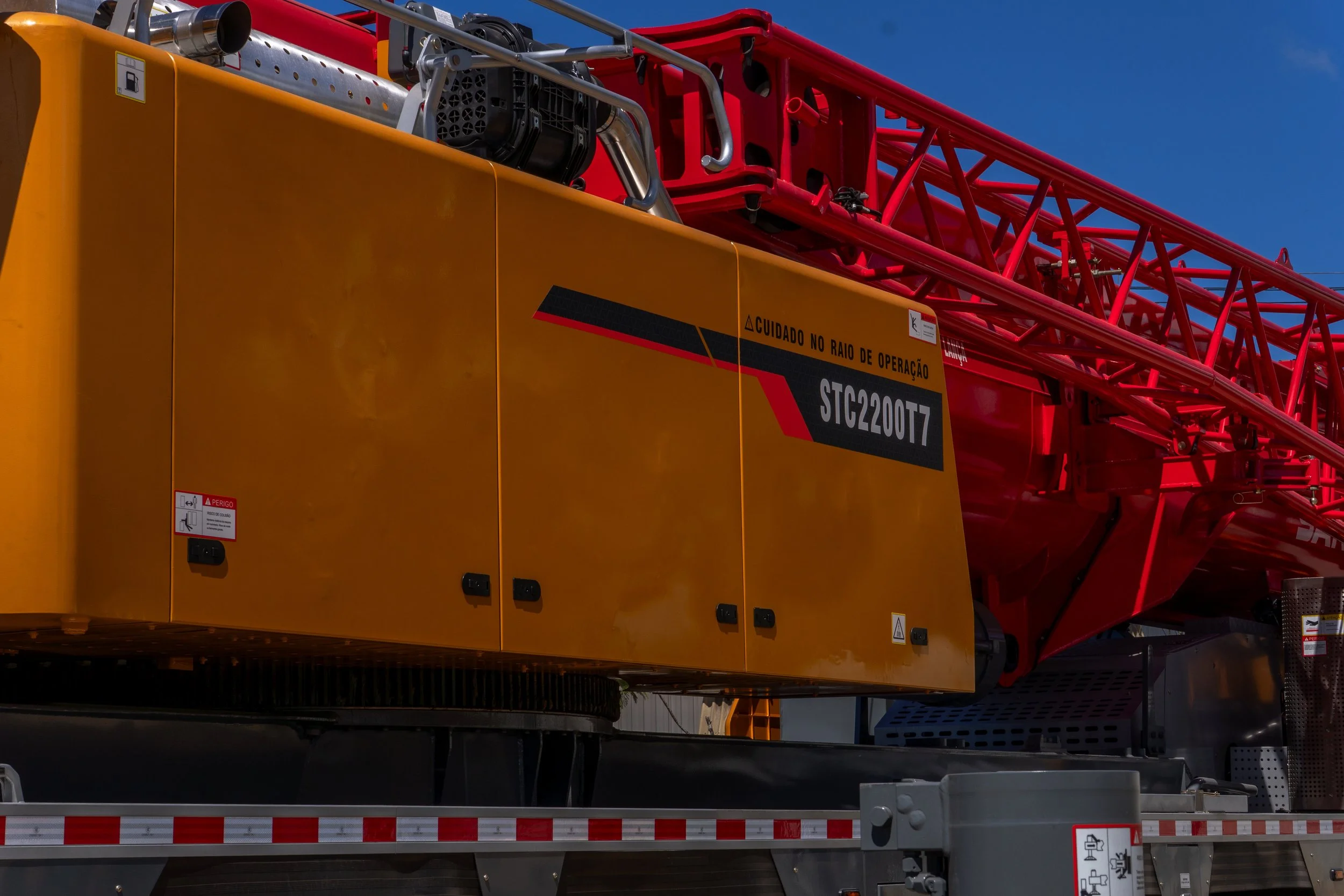 Parte de uma torre de guindaste com componentes amarelos e vermelhos, incluindo uma cabine amarela, estrutura truss tubular vermelha, e um fundo de céu azul.