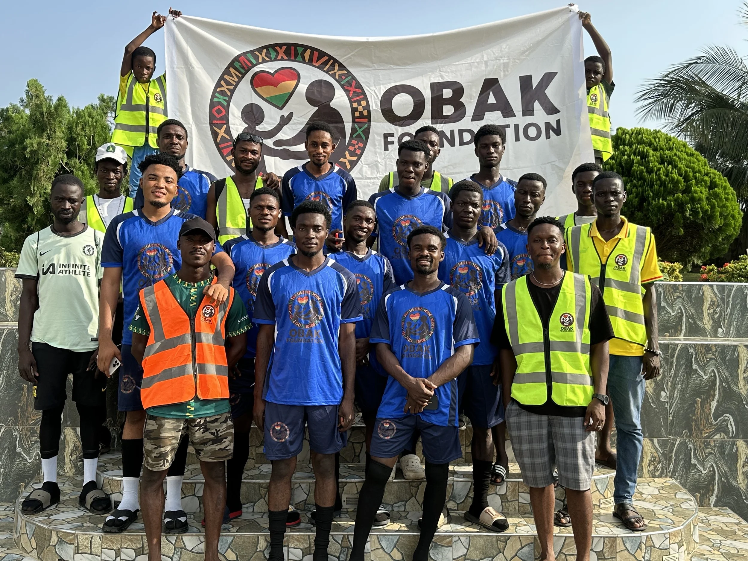 Group of young men, some in blue sports jerseys and others in yellow safety vests, posing outdoors with a banner reading 'OBAK Foundation.' Two children hold the banner at the top, surrounded by lush green trees under a clear sky.
