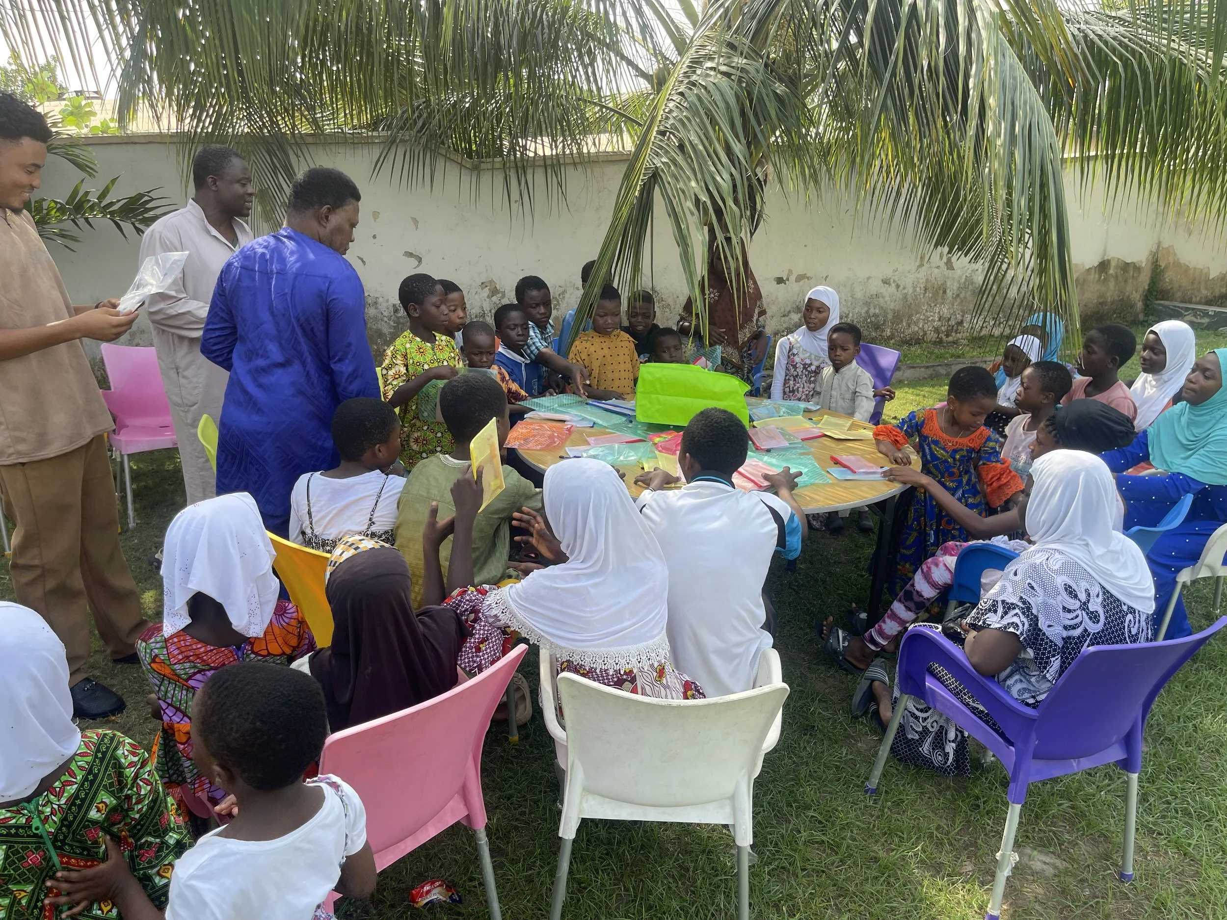 A group of children and adults gathered outdoors around a table with various items, under a palm tree, possibly at a community or school event.