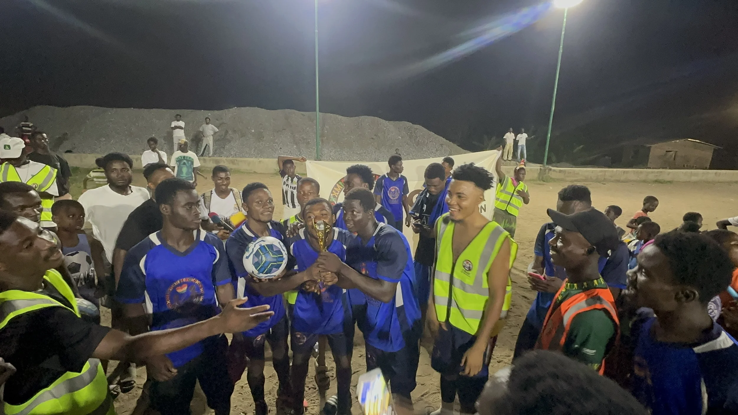 Soccer players celebrating with a trophy and ball at night, surrounded by spectators and officials, on a dirt field.