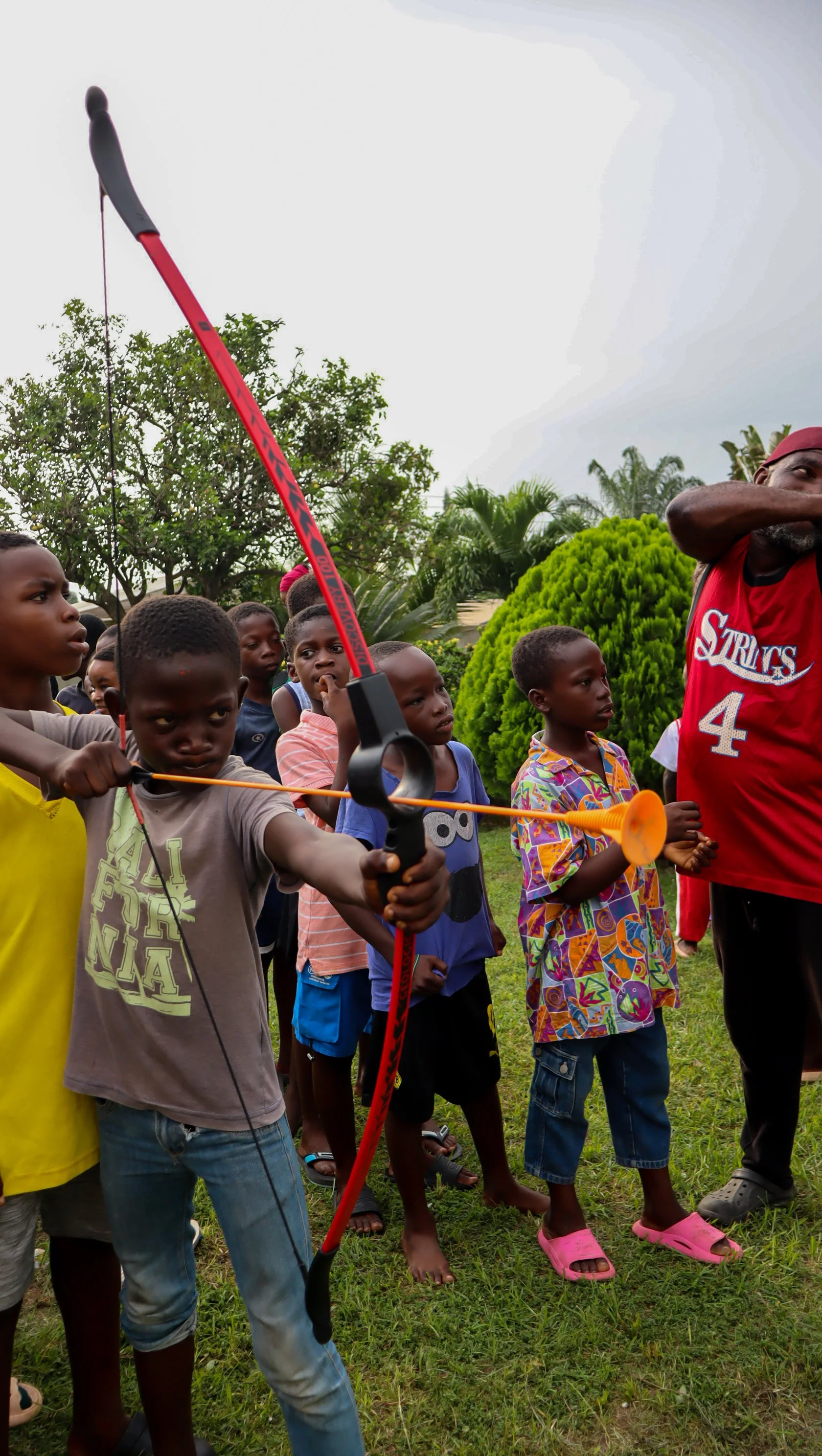 A young boy aiming a bow and arrow as part of a group of children watching outdoors in a grassy area with trees.