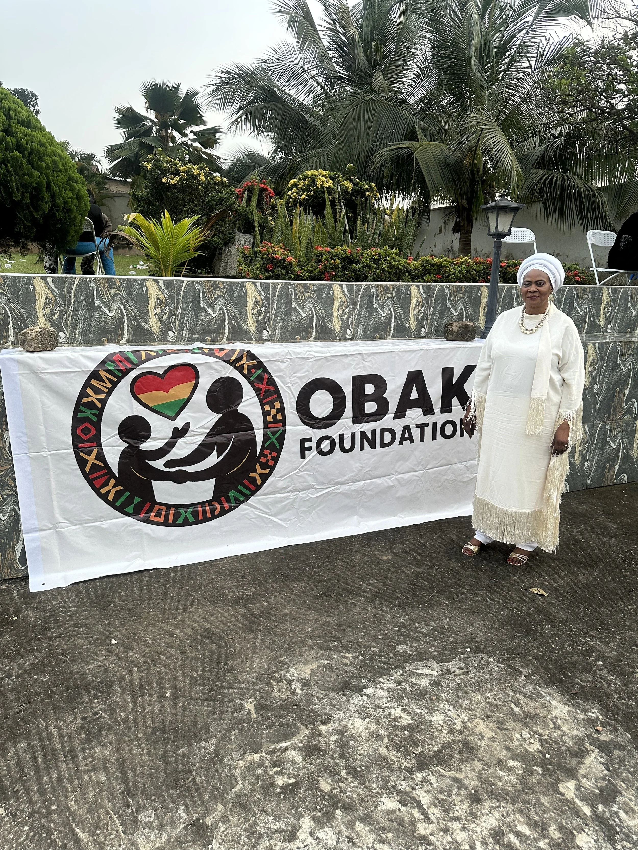 Woman in white traditional dress and headwrap standing next to a banner for the OVAK Foundation outdoors with trees and plants in the background.