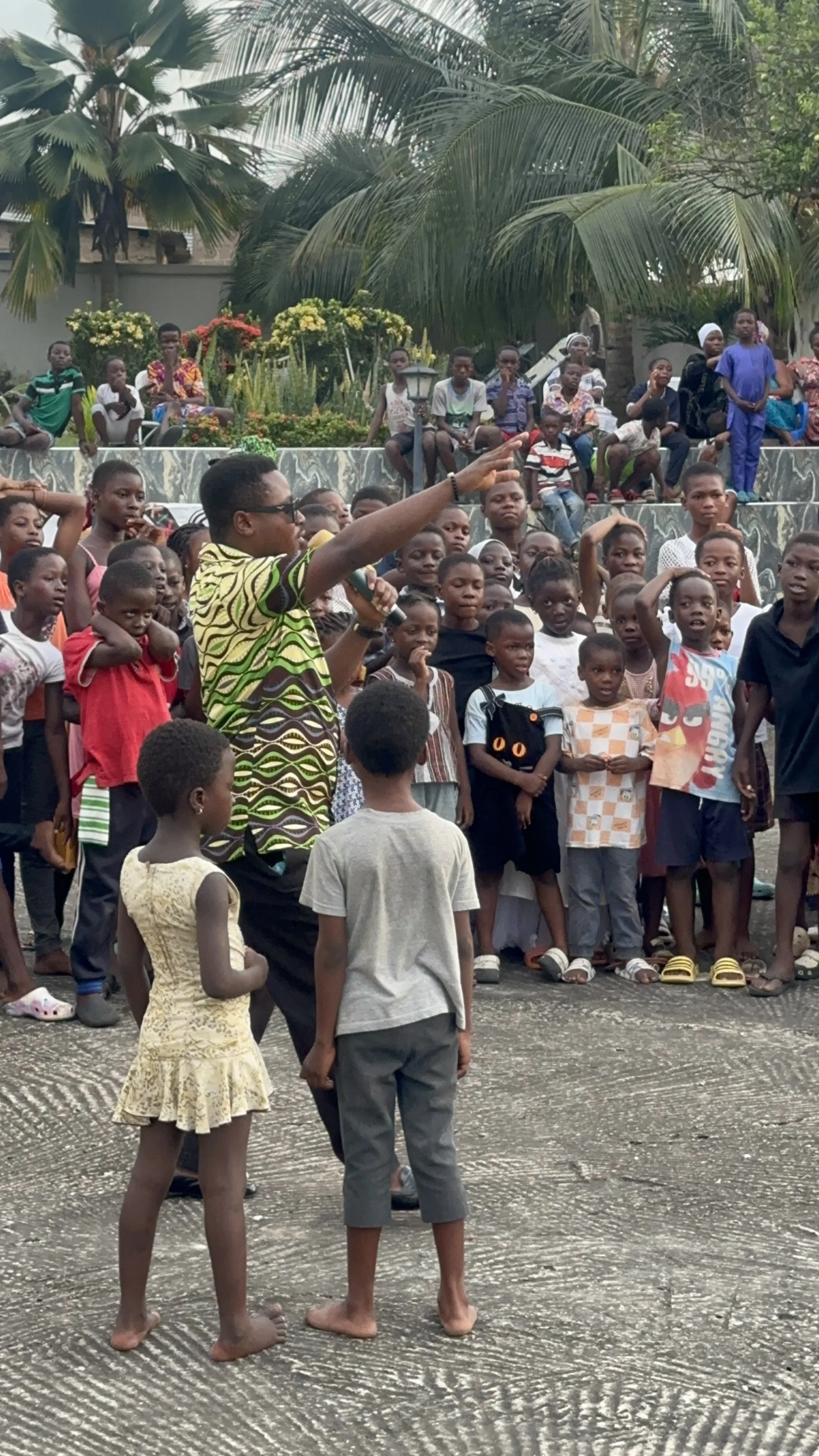 A man speaking into a microphone and gesturing with his hand to a large group of children and teenagers gathered outdoors in a park with palm trees and plants in the background.