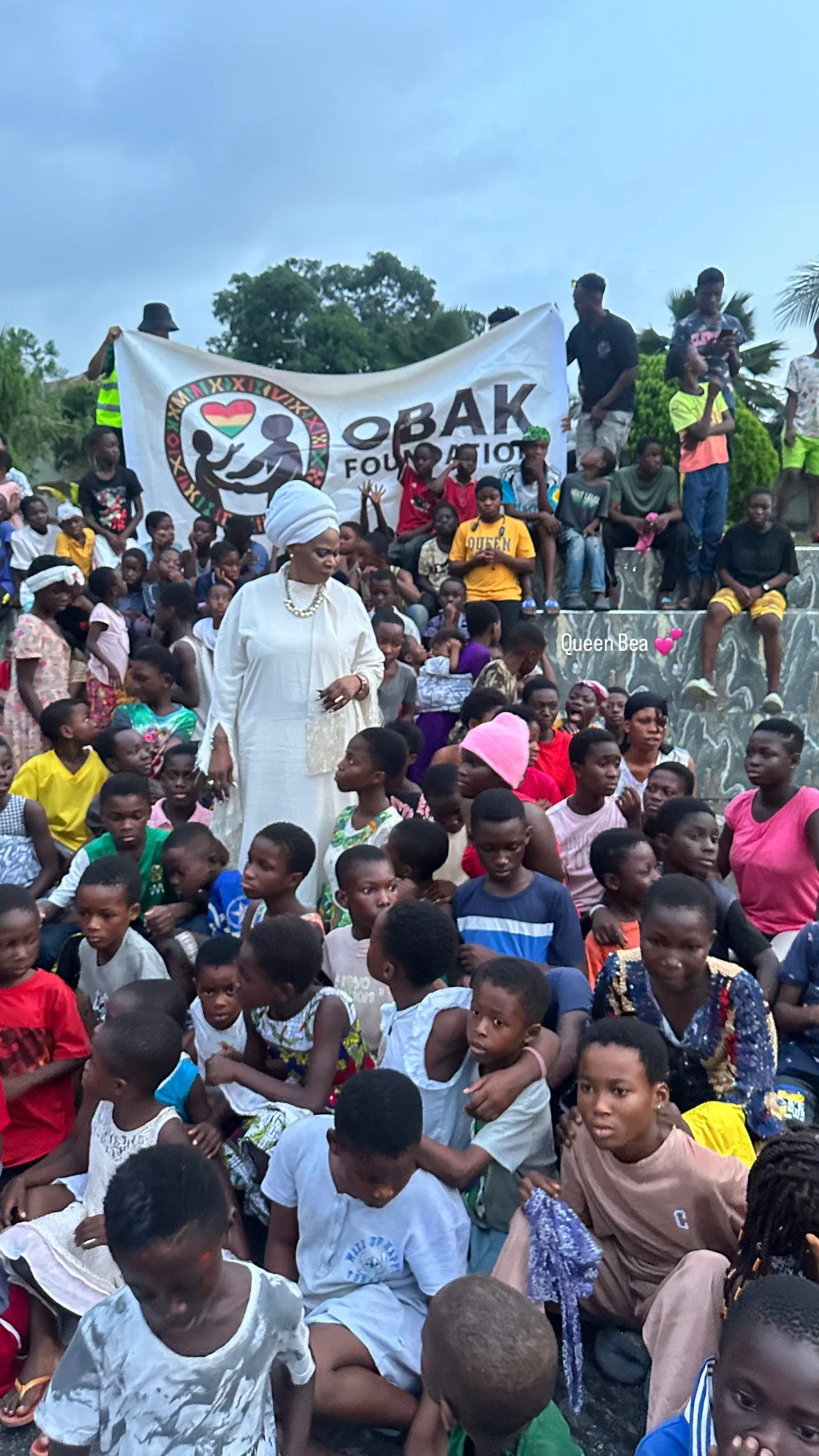 A large group of children and adults gathered outdoors for a community event, with a woman in white clothing and headwrap, and a banner reading 'OBAK FOUNDATION' in the background.