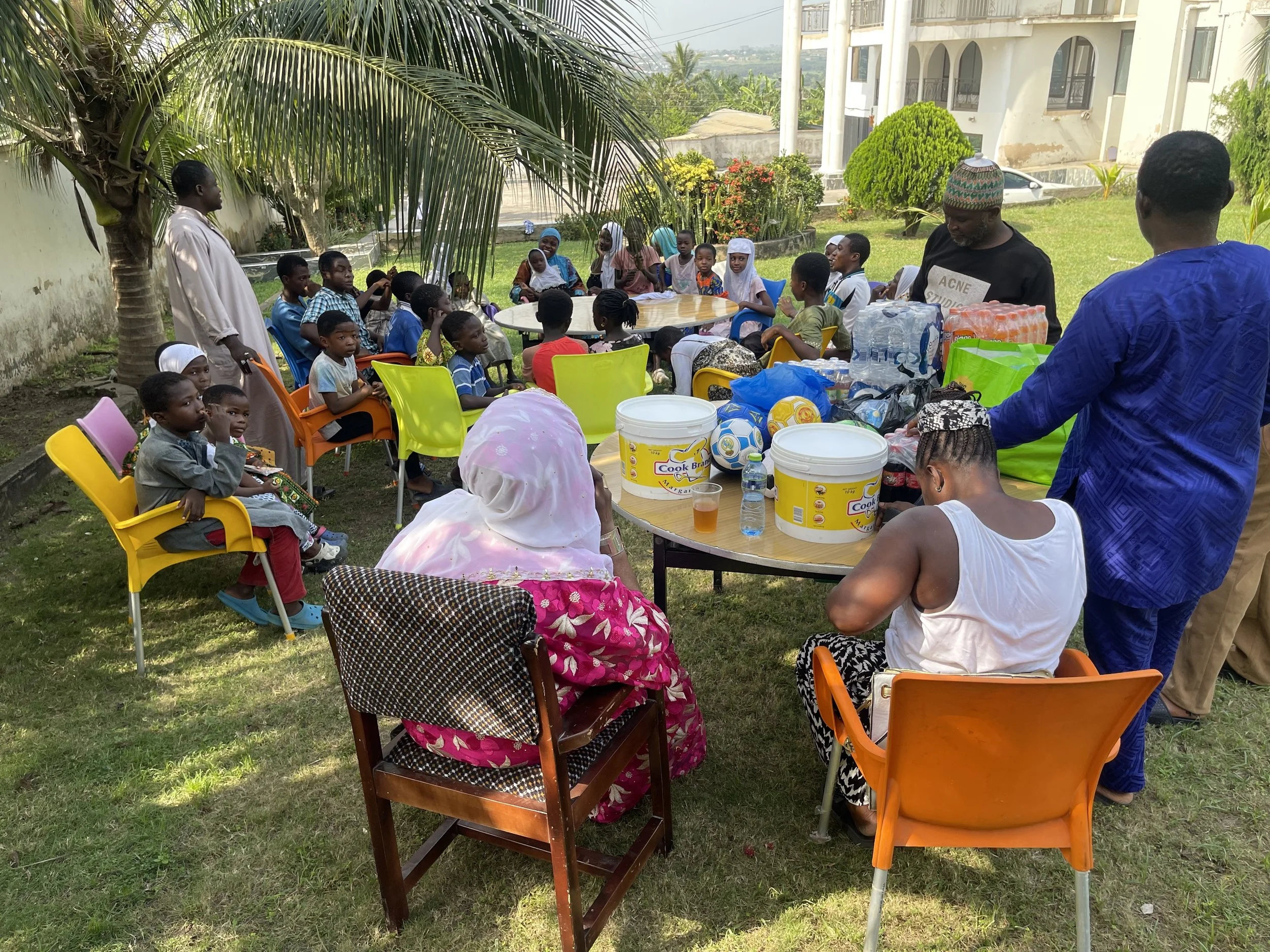An outdoor gathering with children and adults seated around a table with food and drinks, under a tree in a garden near a building.