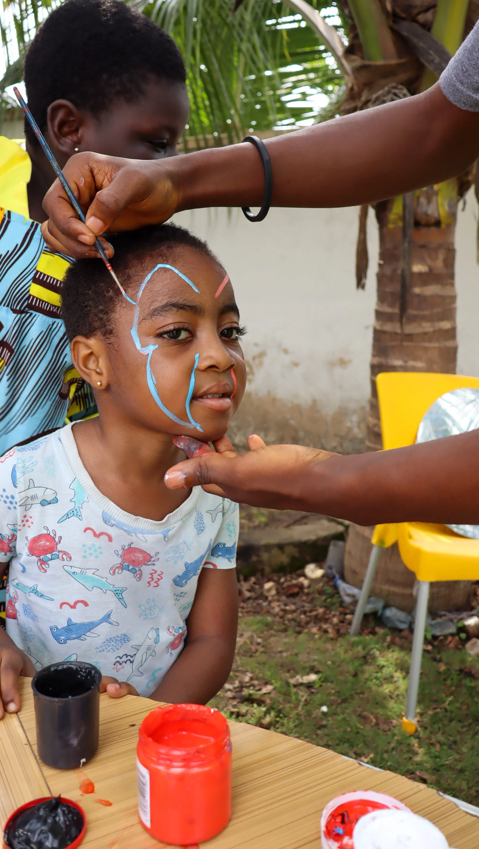 A girl with short curly hair is getting her face painted with blue and red colors by two people. She is sitting at a table with open paint jars in front of her. The background has green foliage, including a palm tree, and outdoor furniture.