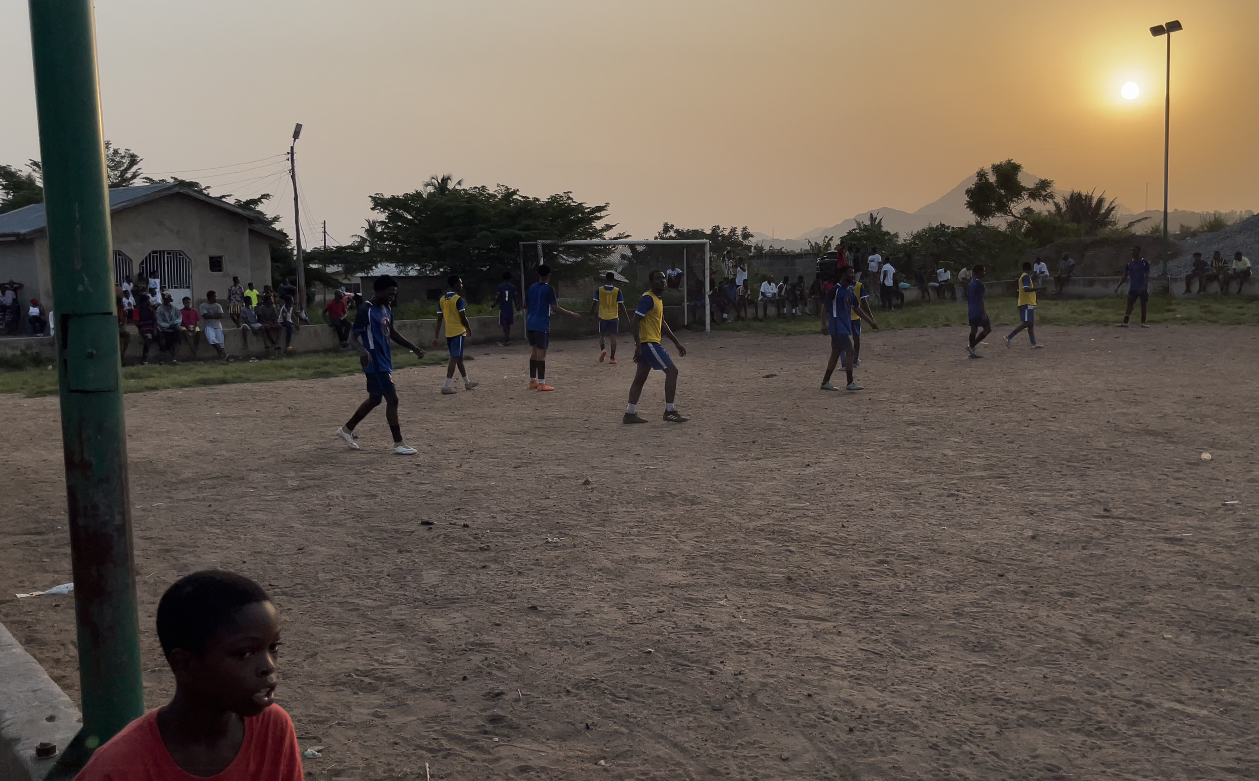 Young boys playing soccer on a dirt field during sunset, with spectators sitting along the sidelines.