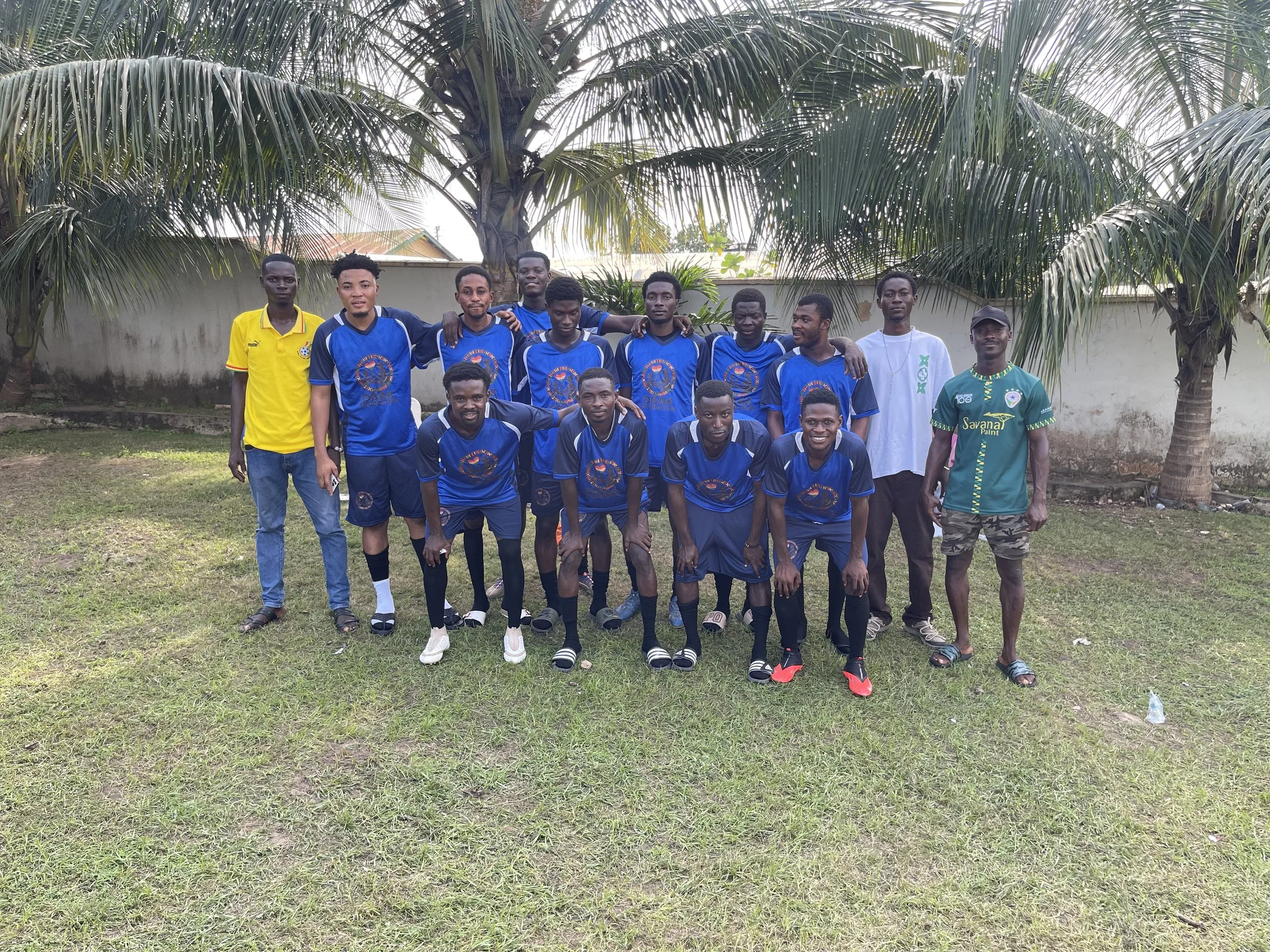 A group of young men in soccer uniforms posing outdoors in a grassy area with palm trees in the background.