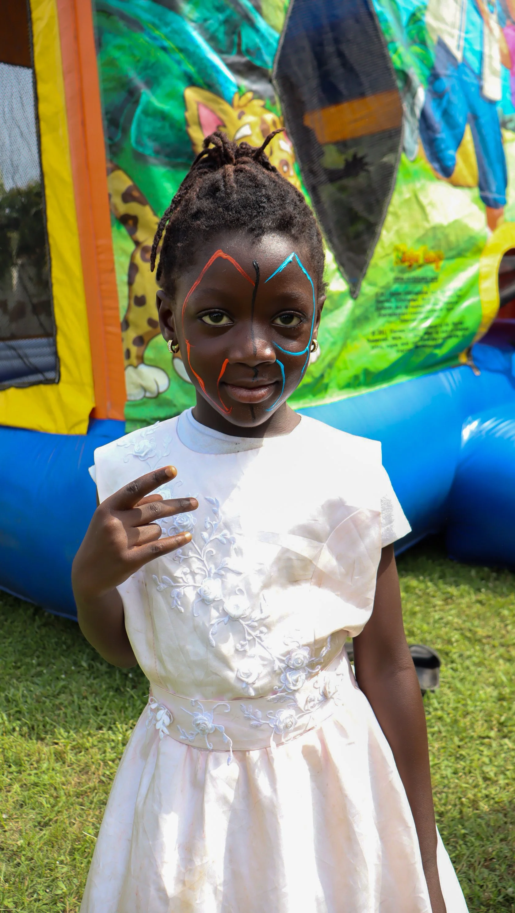 A young girl with face paint stands in front of a colorful bounce house, wearing a white dress with floral embroidery and making a peace sign with her hand.