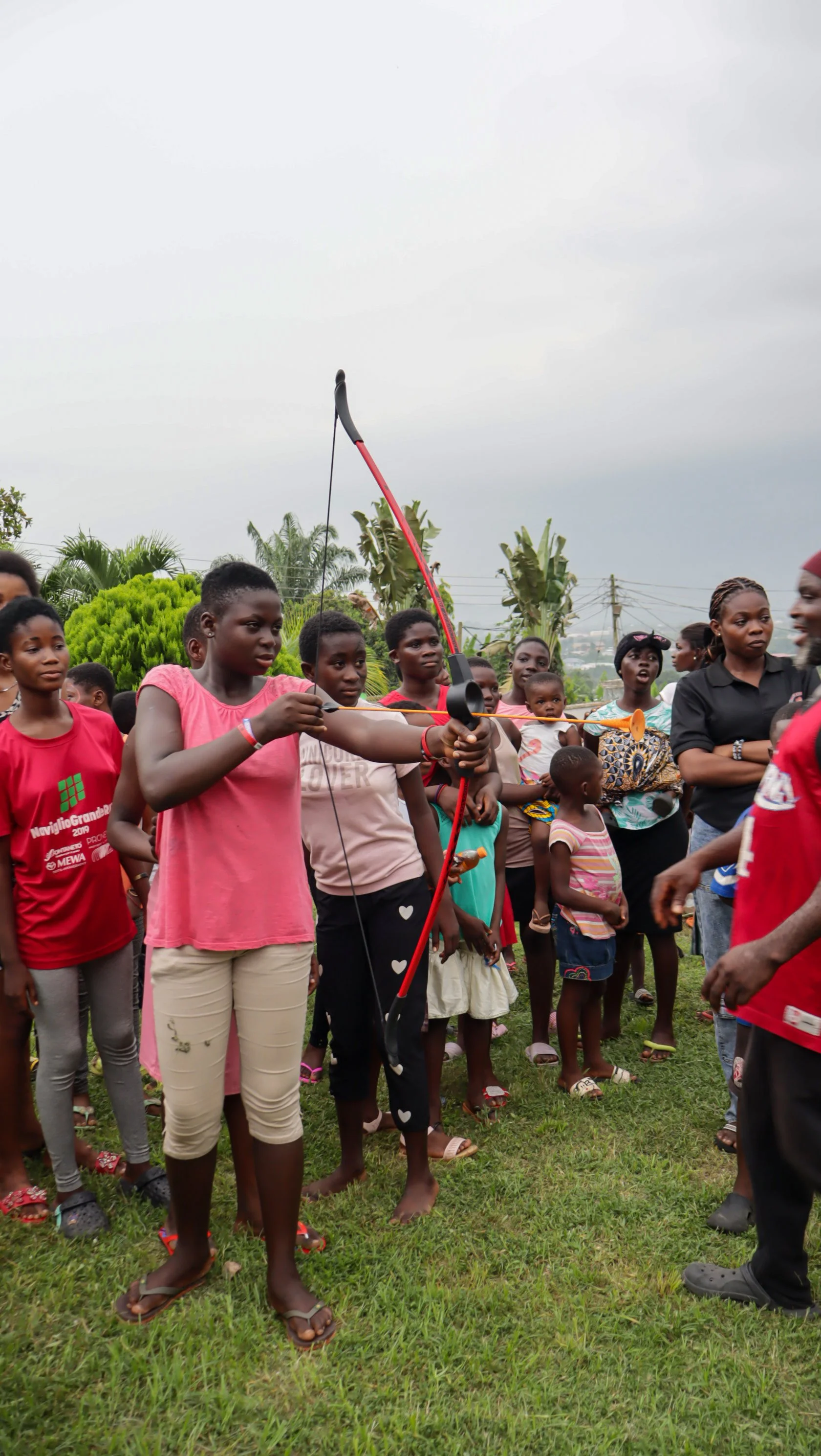 A group of children outdoors, with one girl aiming a bow and arrow, surrounded by other children and adults. There are trees and cloudy sky in the background.
