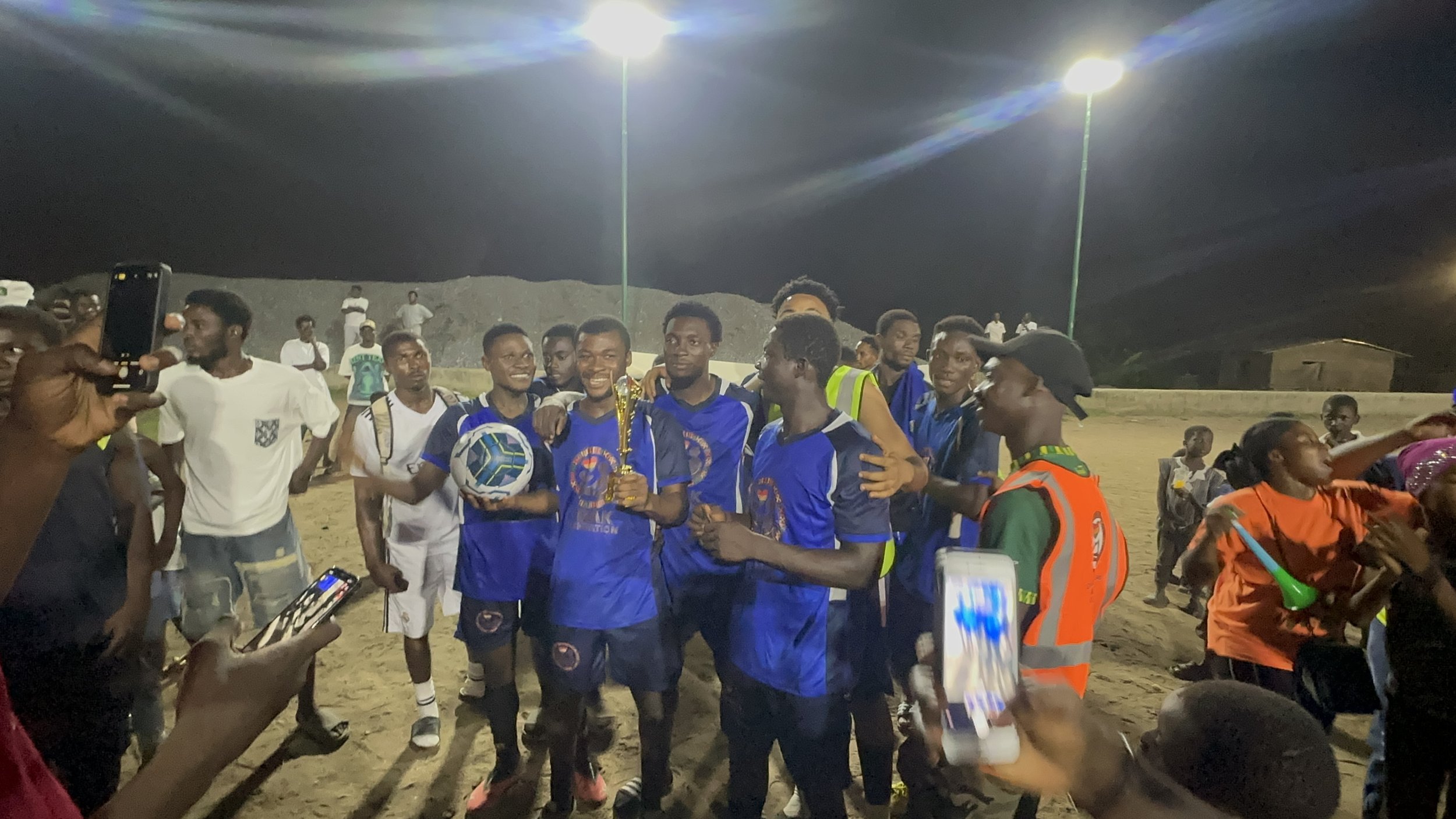 A group of young soccer players on a field at night, celebrating a victory with a trophy, a ball, and smiling. People around are taking photos and cheering.