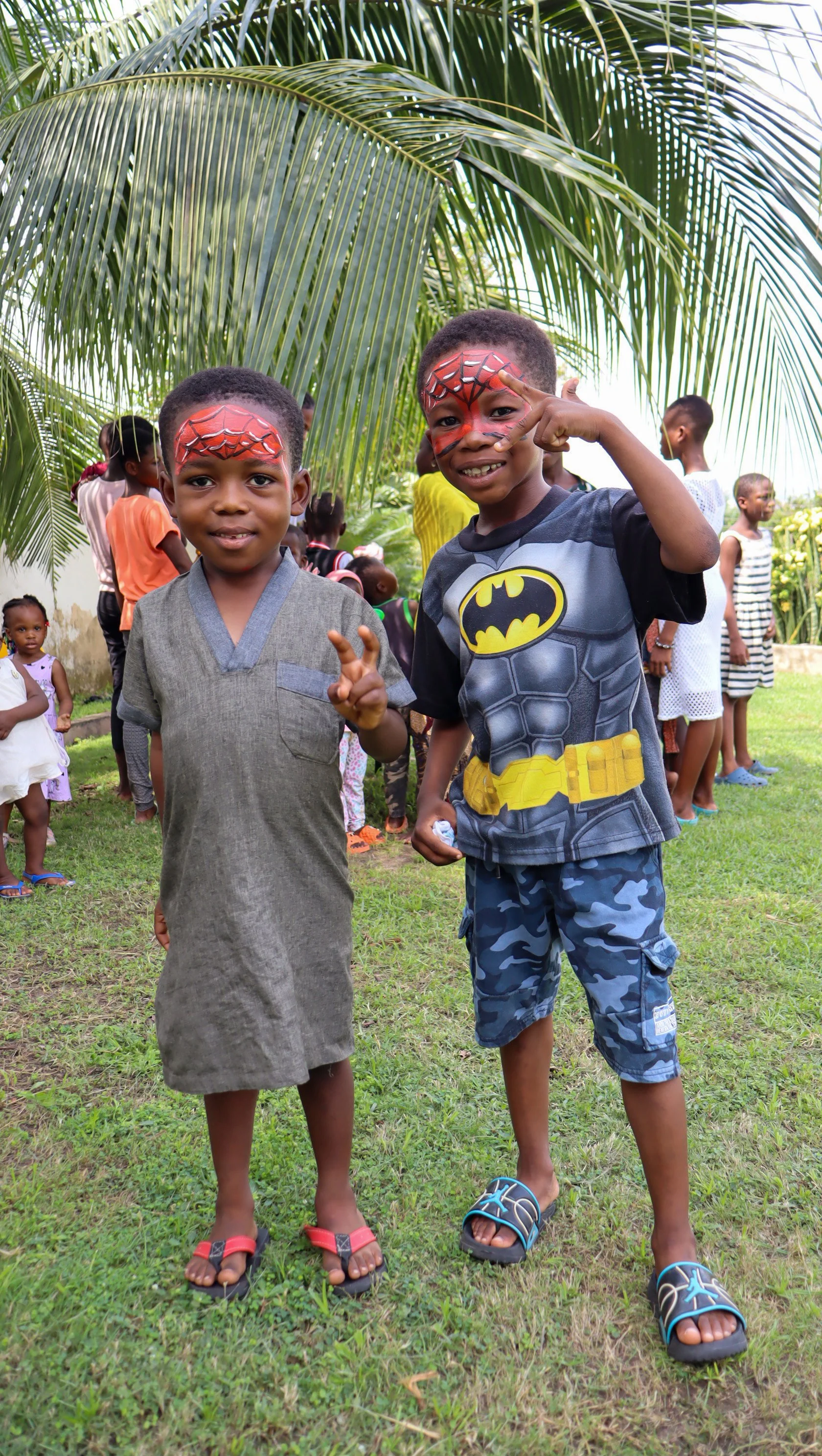 Two young boys with painted faces in a park, facing the camera, surrounded by other children, with large palm trees overhead.