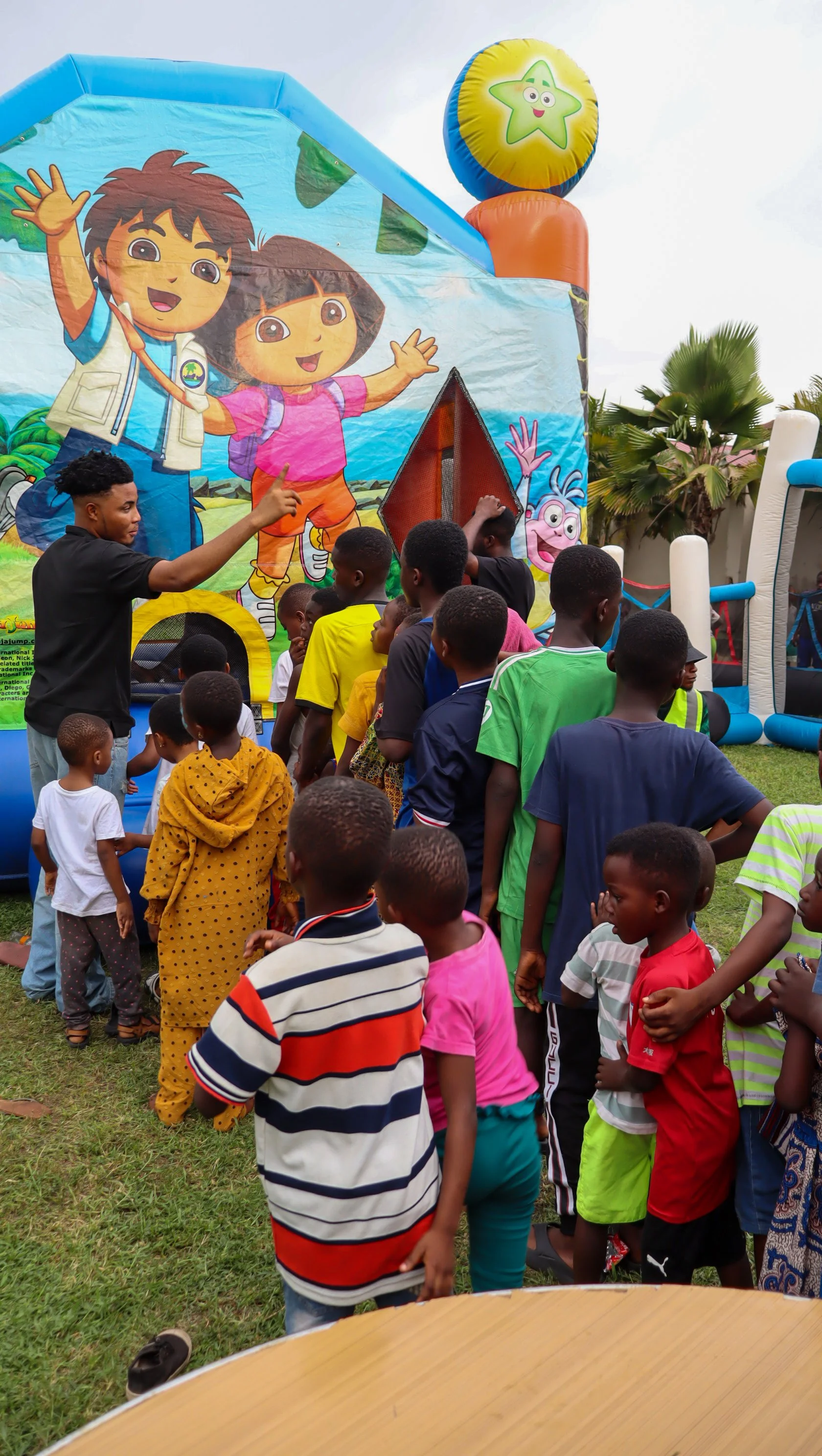 Children, mostly boys, are standing in line outdoors in front of a colorful bouncy castle with cartoon characters, while an adult man is giving instructions or hosting the event.