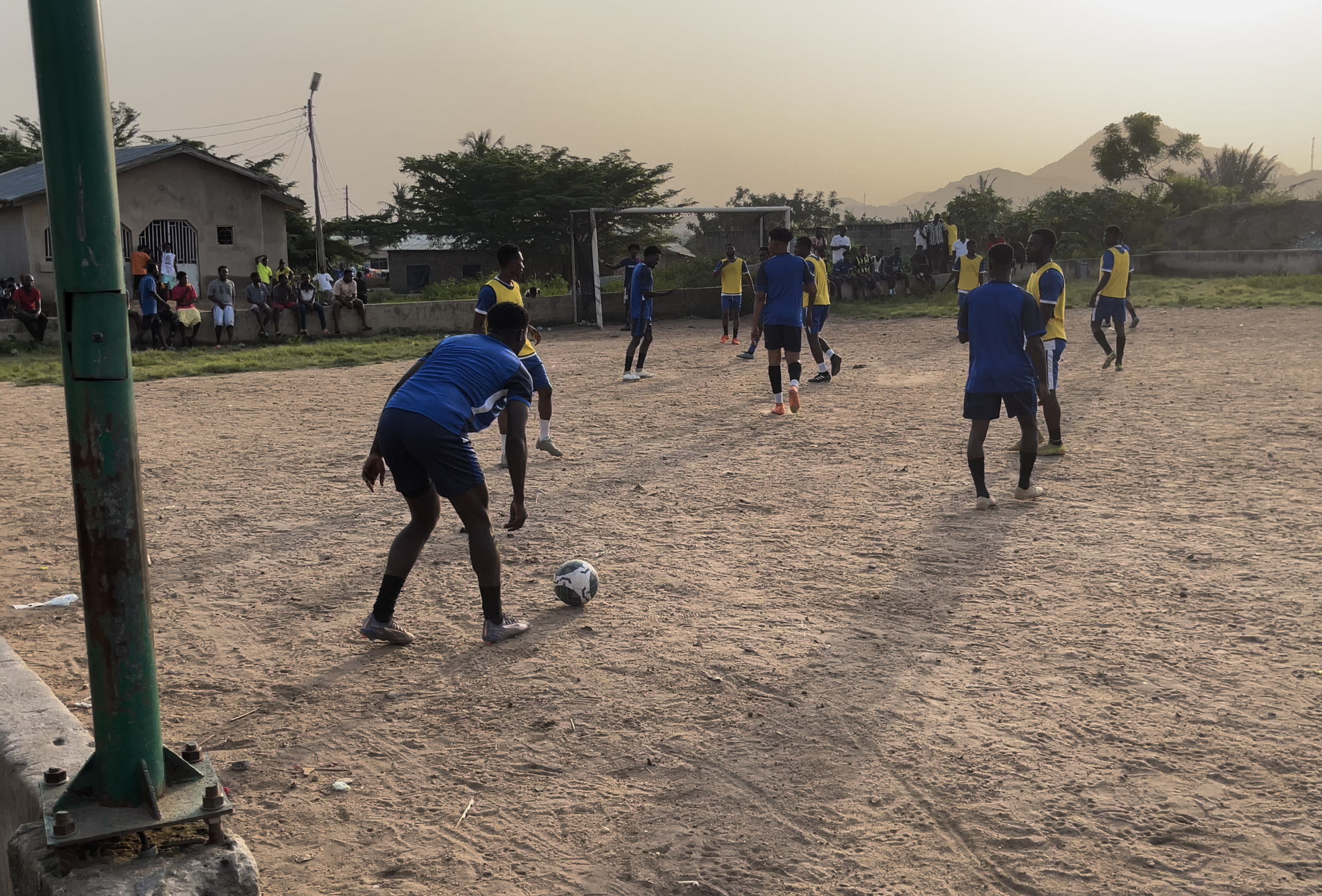Group of young men playing soccer on a dirt field, with some spectators sitting on the sidelines and mountains in the background.