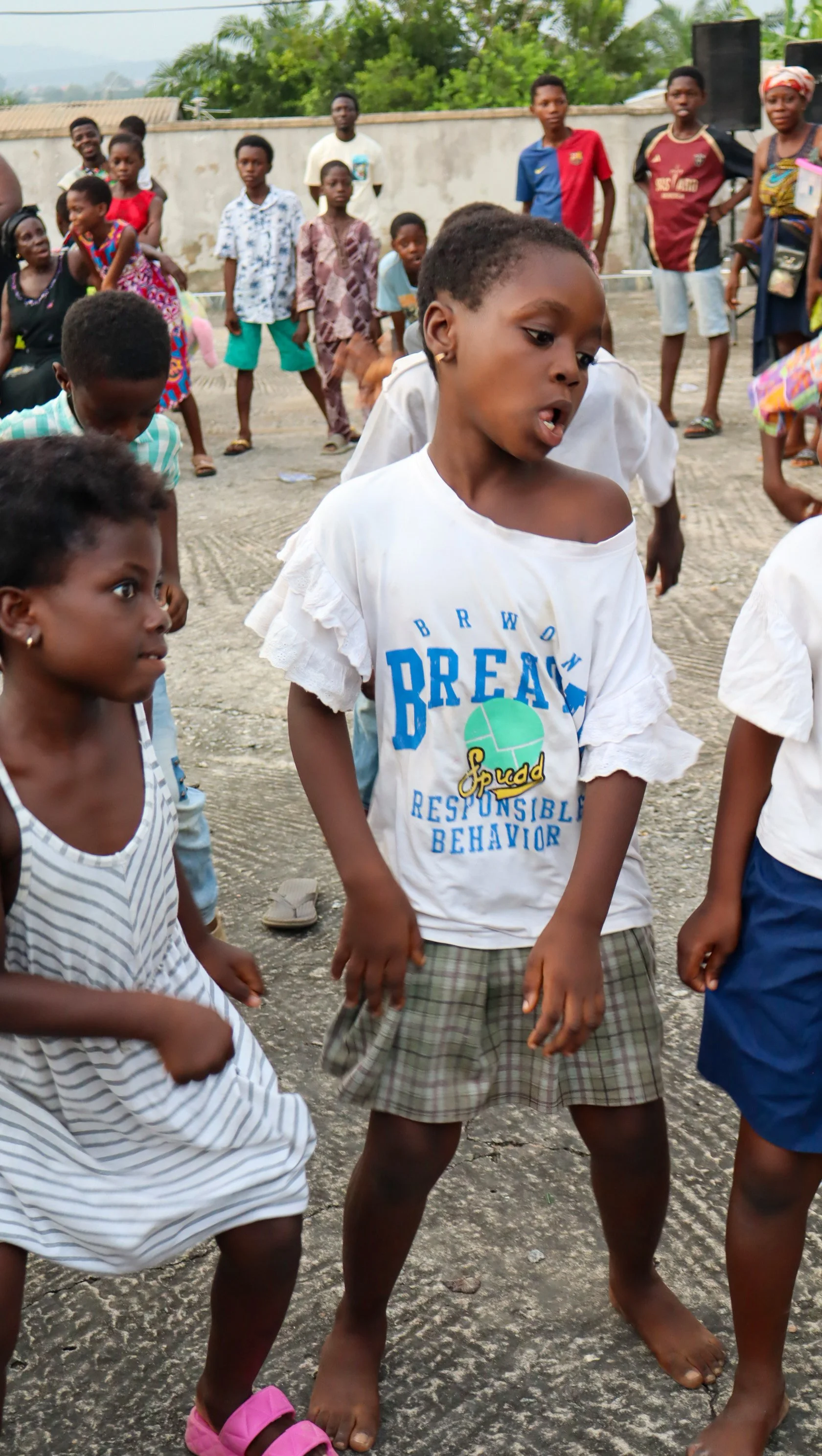 Children dancing outdoors in a group on a rough ground, some with excited expressions, with more people in the background near a wall and trees.