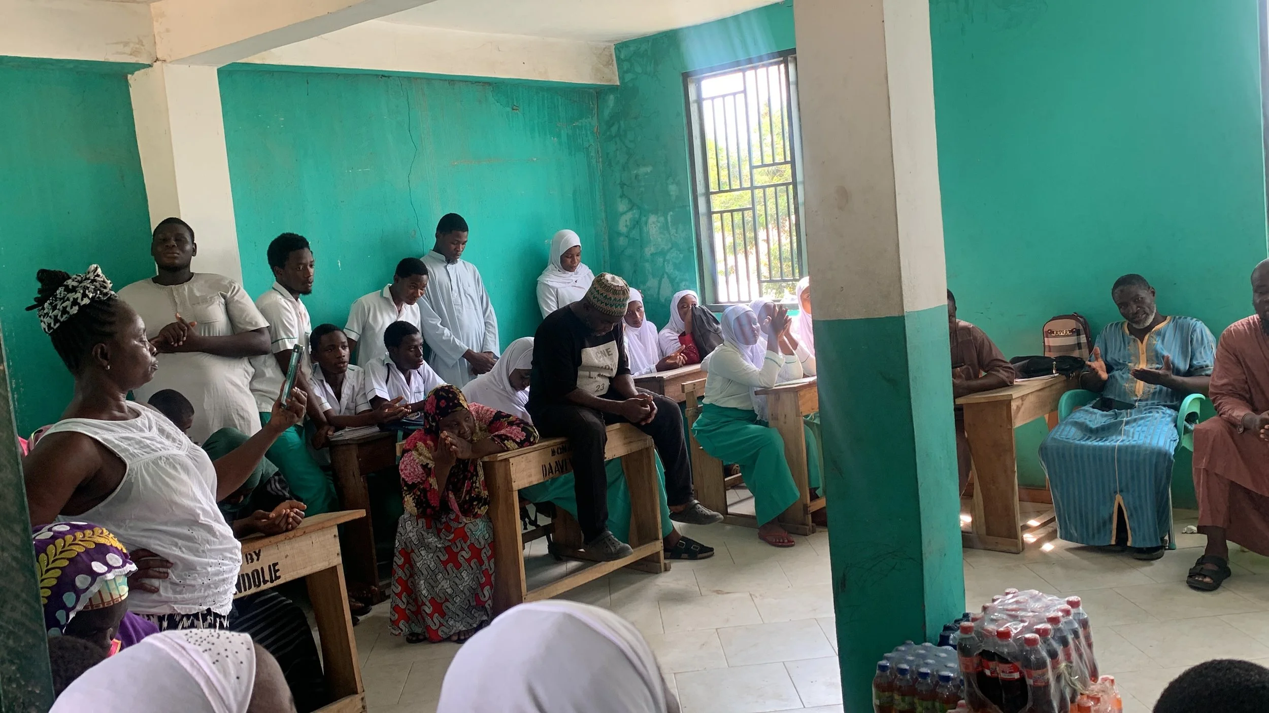 Group of people gathered in a room with green walls, sitting on benches, some wearing traditional clothing, while a woman in the front takes a photo or video with her phone. A man in blue traditional attire appears to be speaking or explaining someth