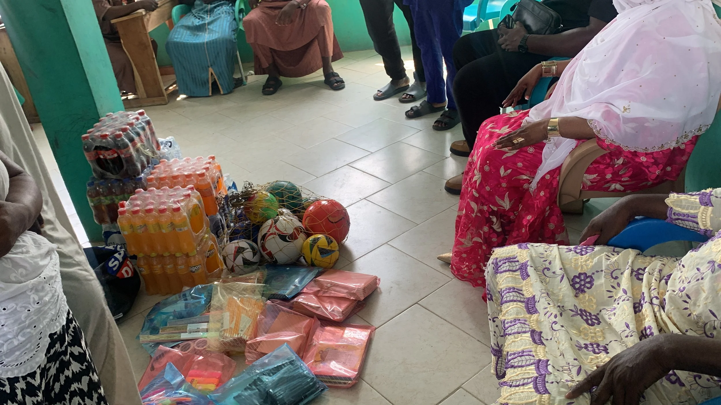 People seated in a circle, with supplies and gifts placed on the floor in the center, including bottled beverages, balls, and wrapped packages, in a room with colorful walls and tiled floor.