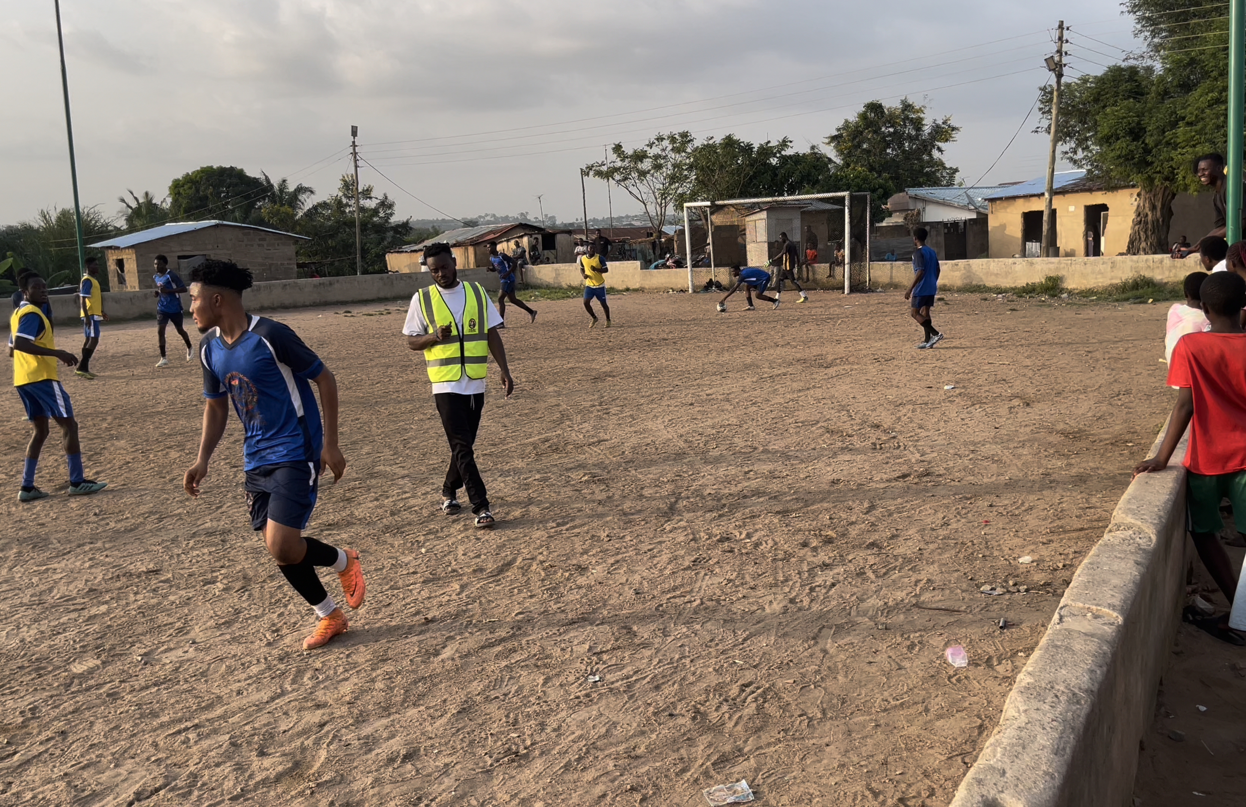 Young boys playing soccer on a dirt field with makeshift goalposts, some spectators watching from the sidelines, in a rural area with small houses and trees in the background.
