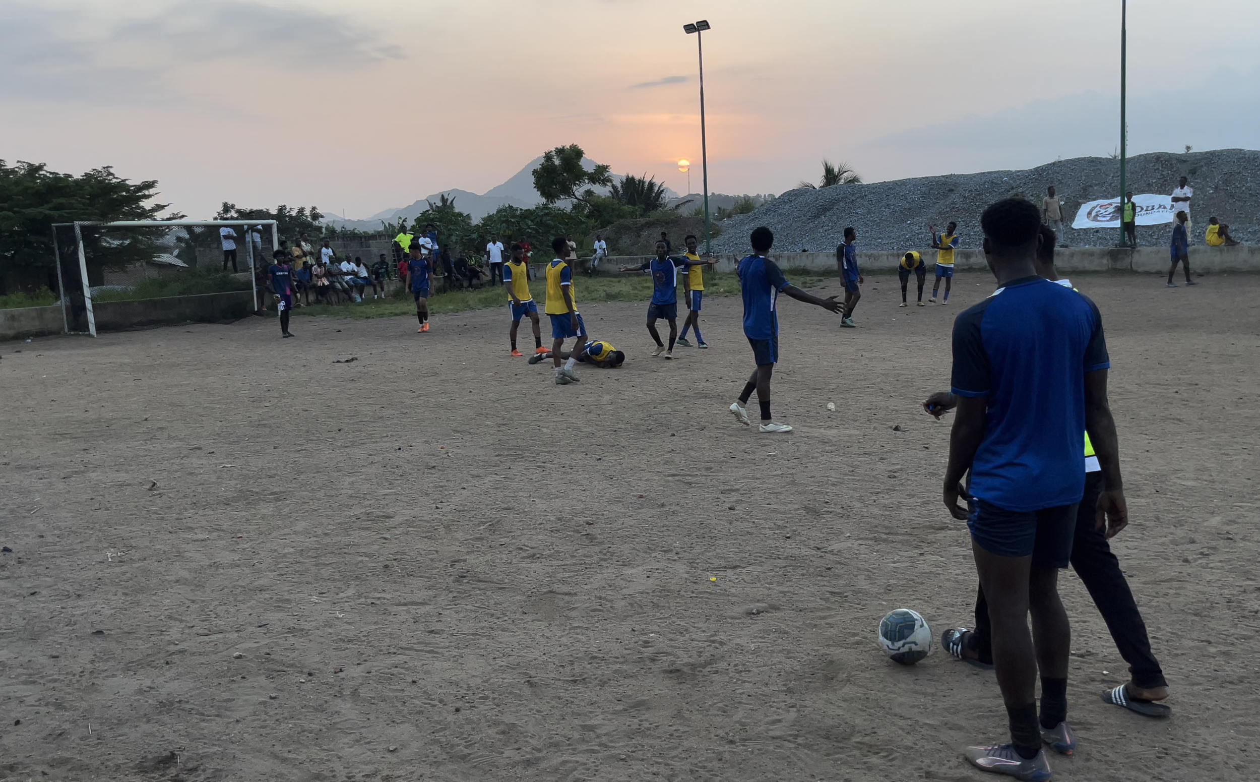 Players on a dirt soccer field during sunset, some wearing yellow and blue jerseys, with spectators on the sidelines and a goalpost on the left.