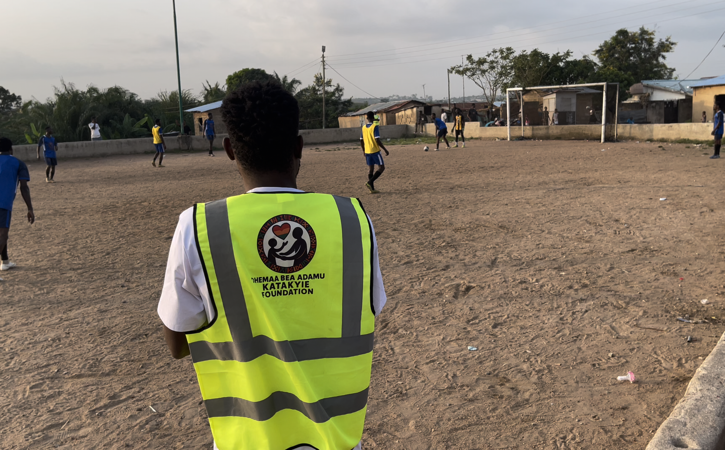 A group of boys playing soccer on a dirt field, with some houses and trees in the background, and a person wearing a yellow vest with a logo and text on the back observing the game.