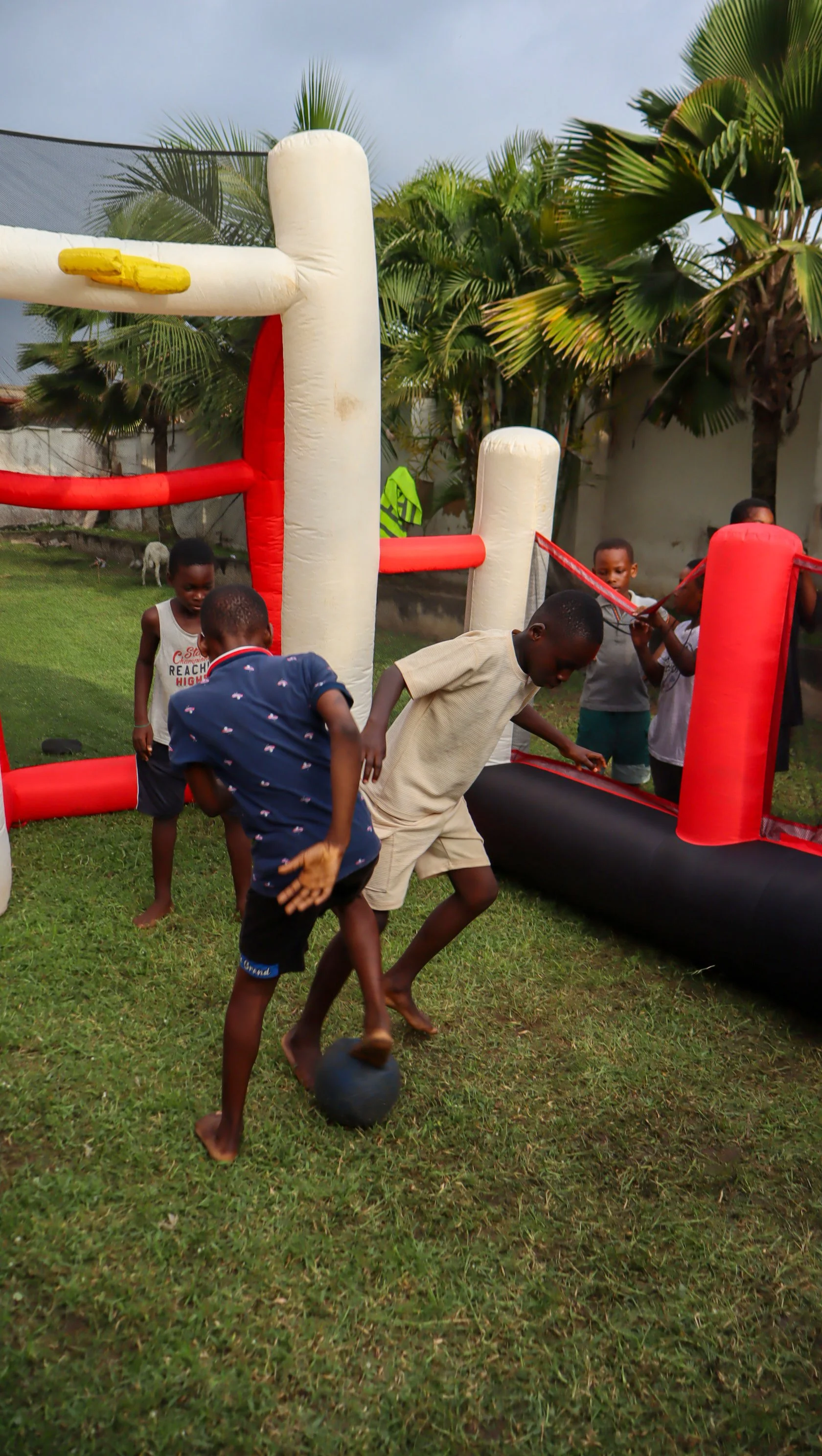 Children playing a game of soccer around an inflatable soccer goal set up outdoors in a yard with grass and palm trees.