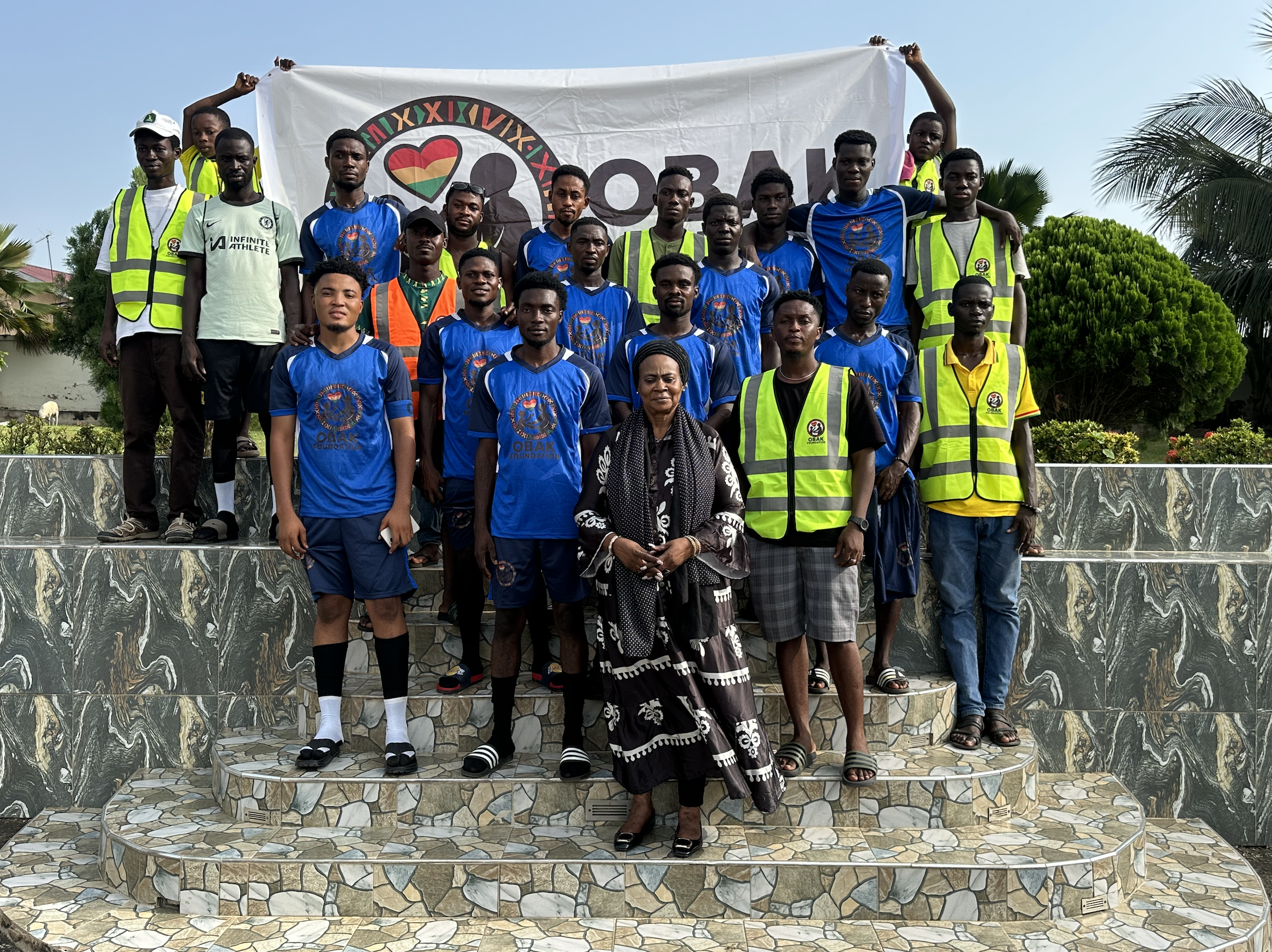 Group photo of young male athletes and officials, some wearing blue sports uniforms and yellow safety vests, standing on steps outdoors with a large banner behind them, with trees and a blue sky in the background.