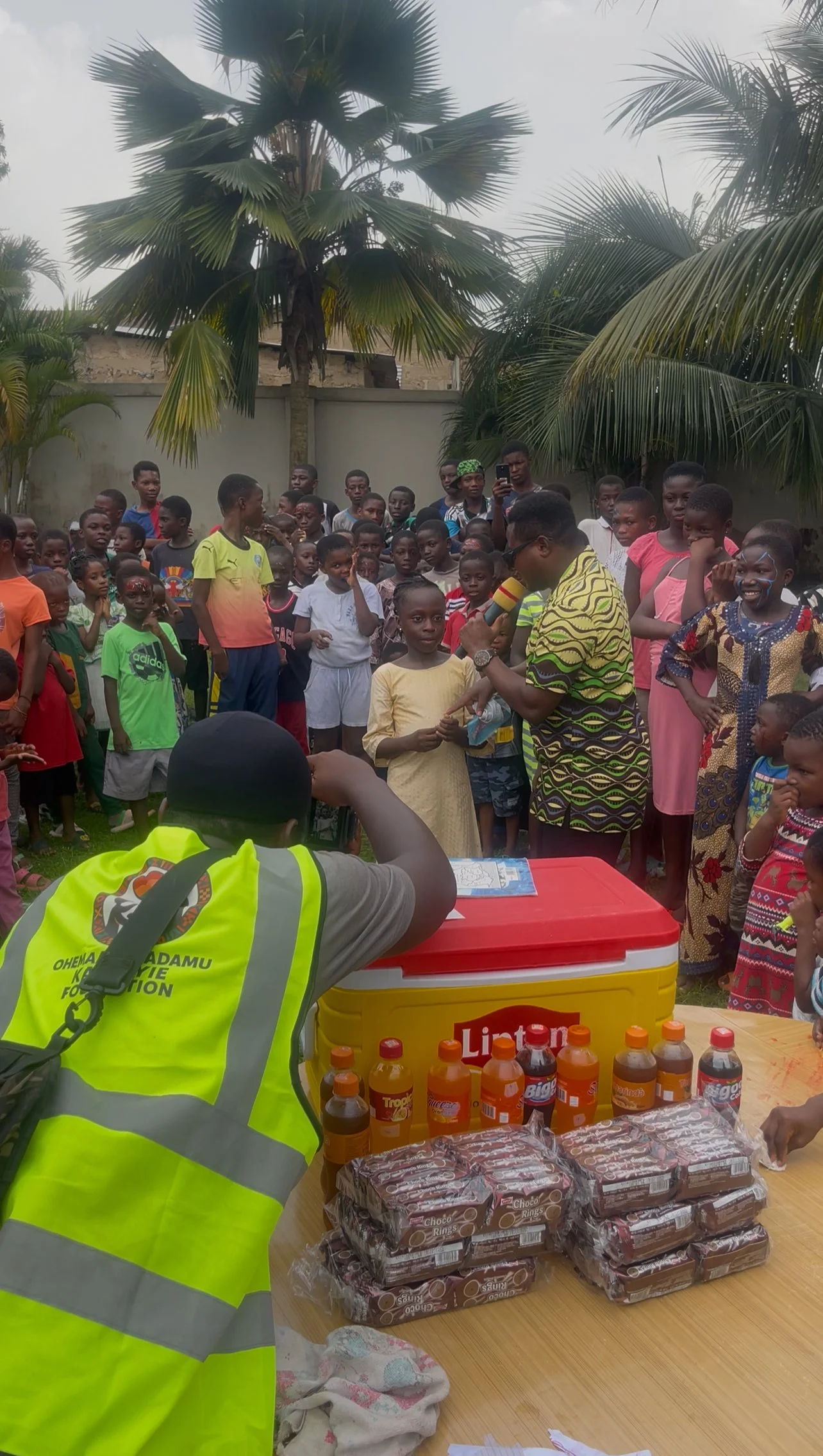 A large group of children and adults gather outdoors around a table with snacks and drinks, as a person in a yellow vest with the text "OHEMA ADAMU KABIRIE FOUNDATION" talks to a girl in yellow. Some children are smiling, while others watch attentive