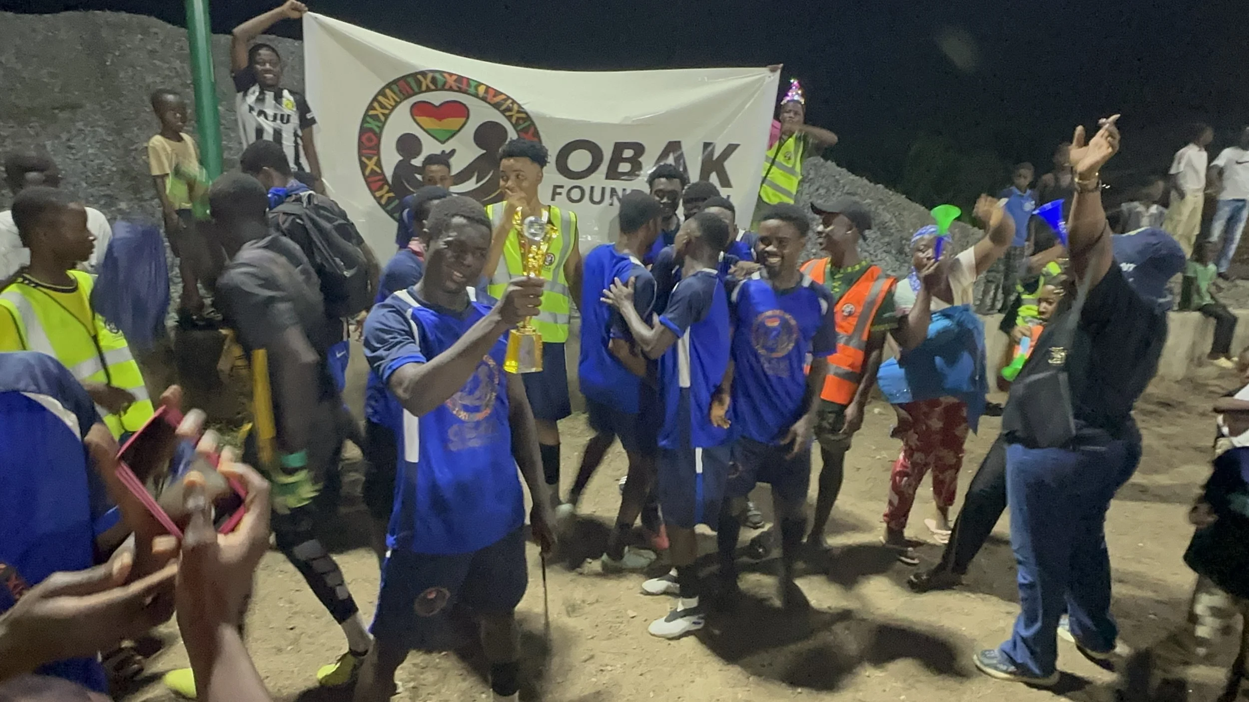 Group of young people celebrating outdoors at night, some holding a trophy, with a large banner reading "OBAK Foundation" and a logo featuring two children and a rainbow heart.