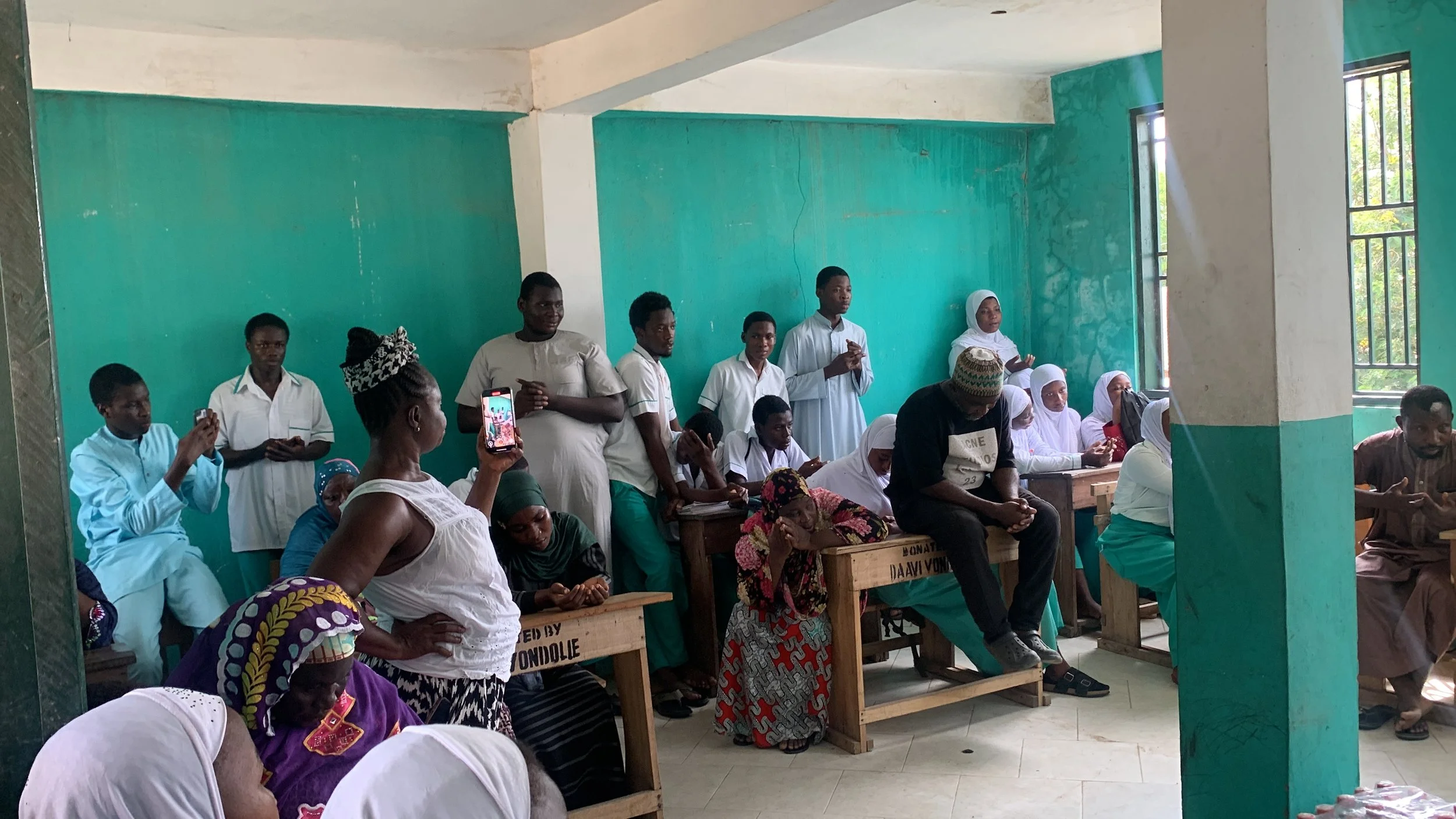 Group of people gathered indoors, some sitting and some standing, in a room with turquoise walls and windows, participating in a meeting or event.