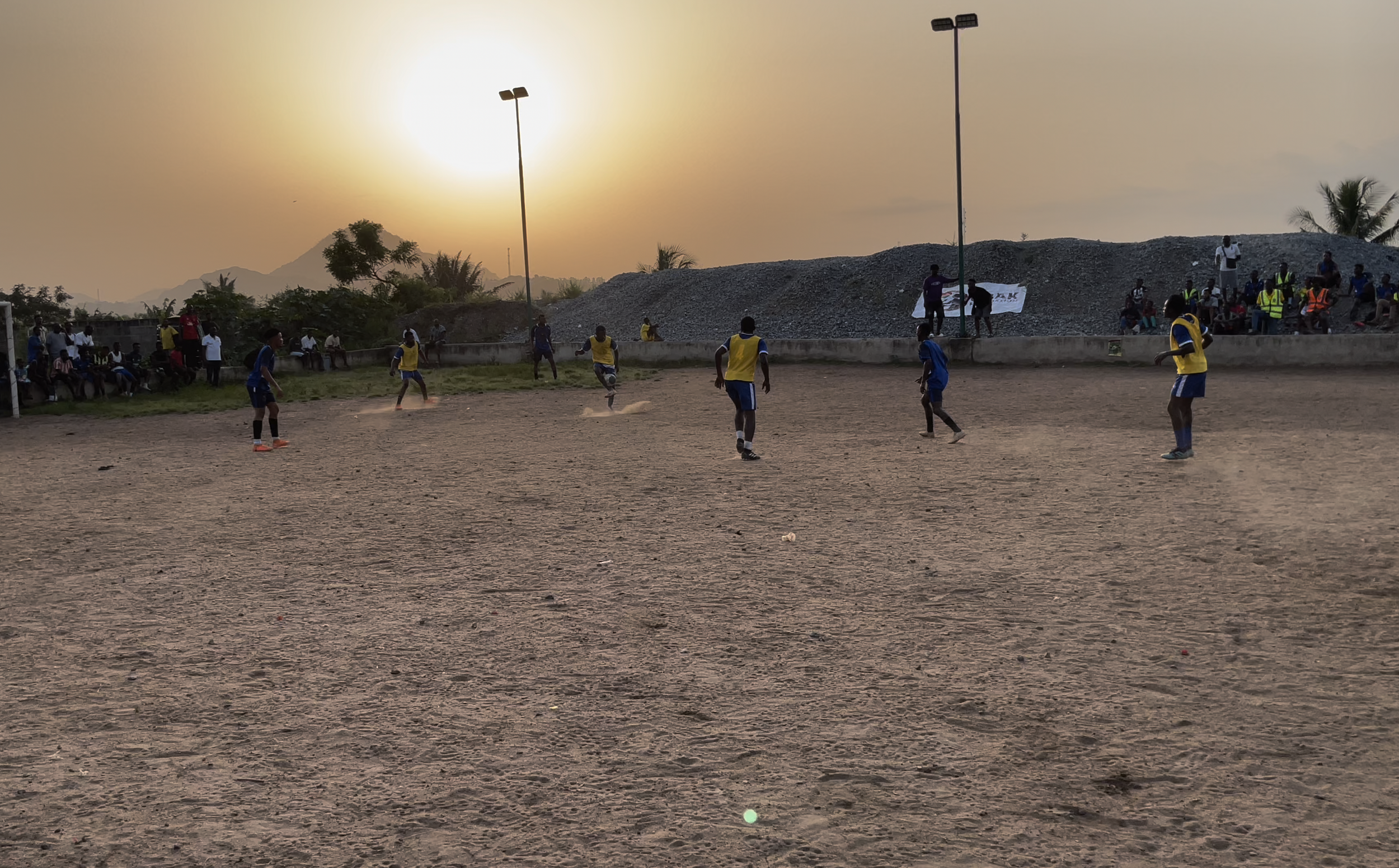 Soccer game taking place on a dirt field during sunset with players wearing blue and yellow jerseys, and spectators watching from the sidelines.