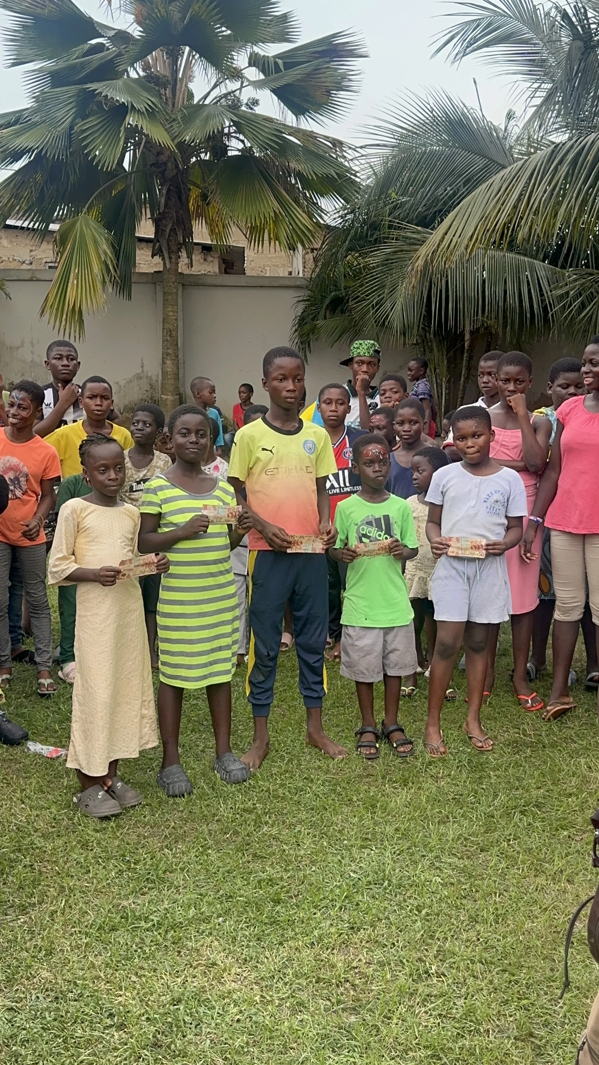 Group of children and a few adults outdoors on grass, some holding certificates, standing under palm trees.