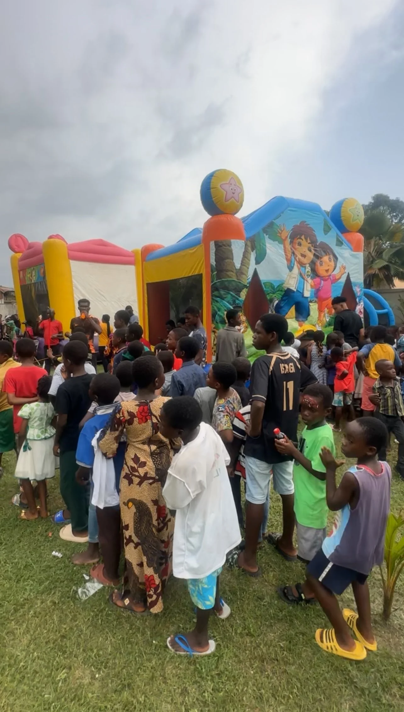People gathered around a colorful inflatable bounce house with cartoon characters and beach ball decorations on a grassy field.