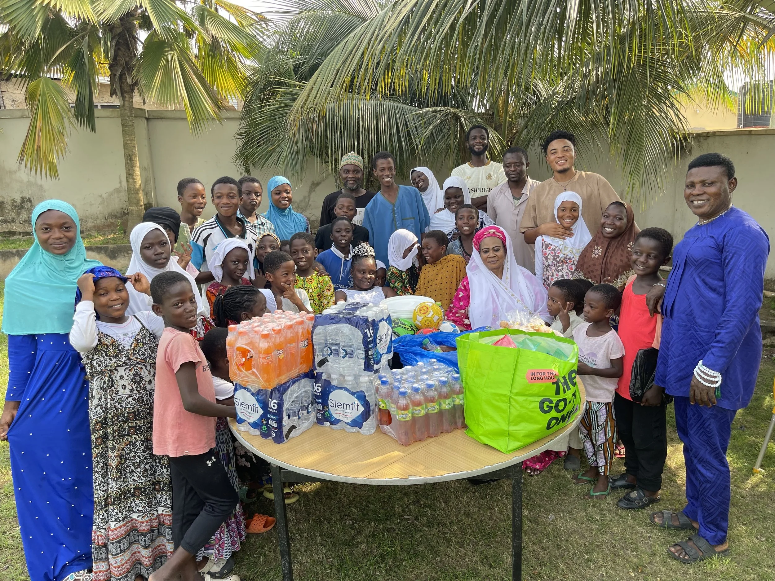 A large group of children and adults gathered outdoors under palm trees, smiling around a table with various food and drinks, celebrating an event.
