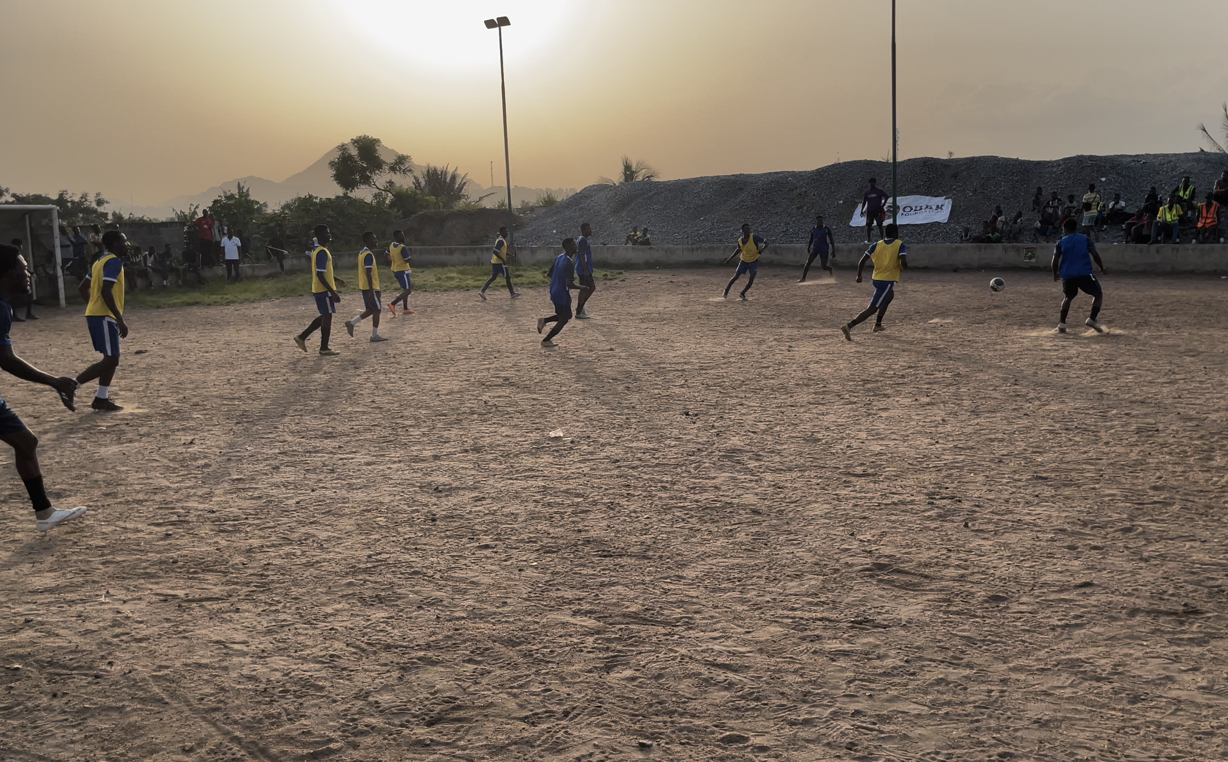 Young boys playing soccer on a dirt field during a sunset, with some spectators and hills in the background.