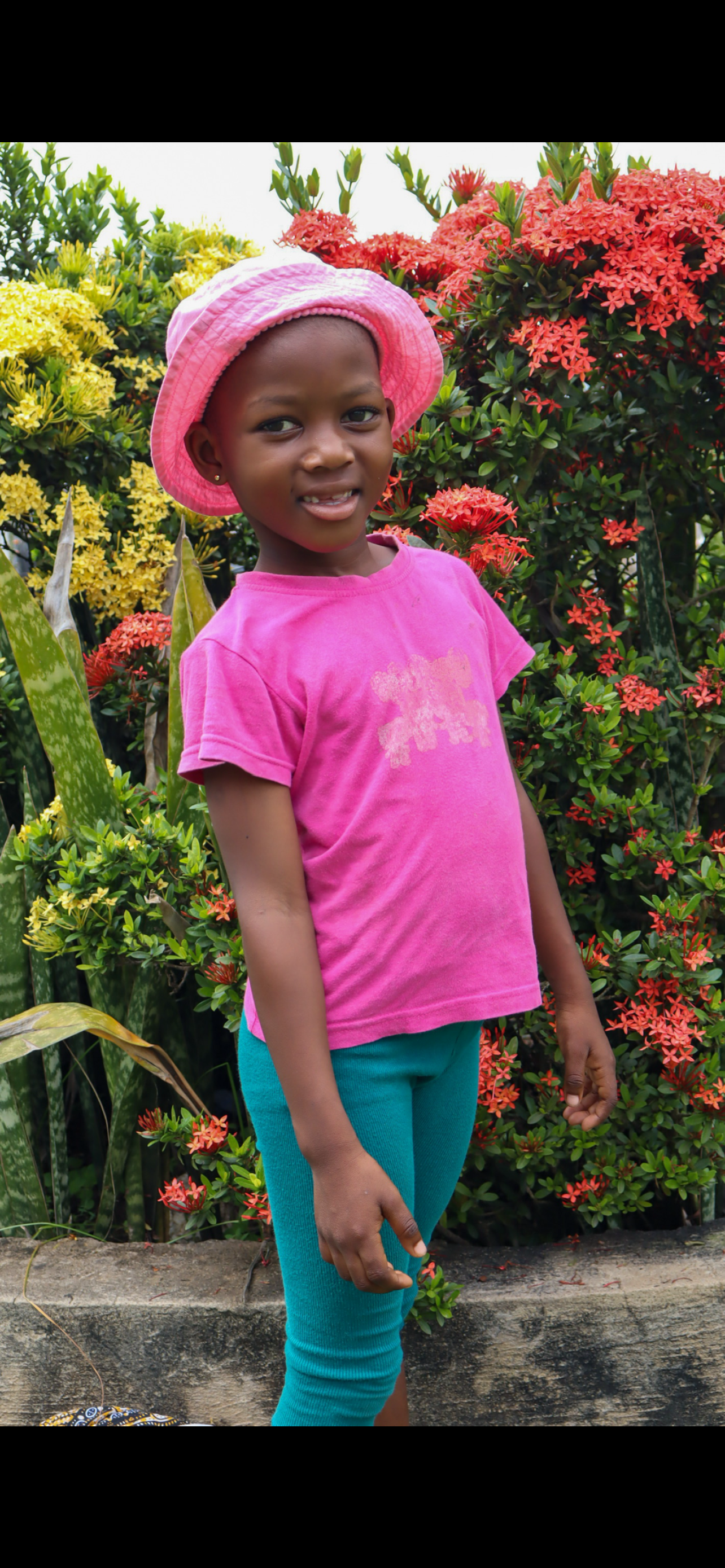 A young girl standing outdoors in front of vibrant red, yellow, and green plants, wearing a pink hat, pink t-shirt, and turquoise pants.