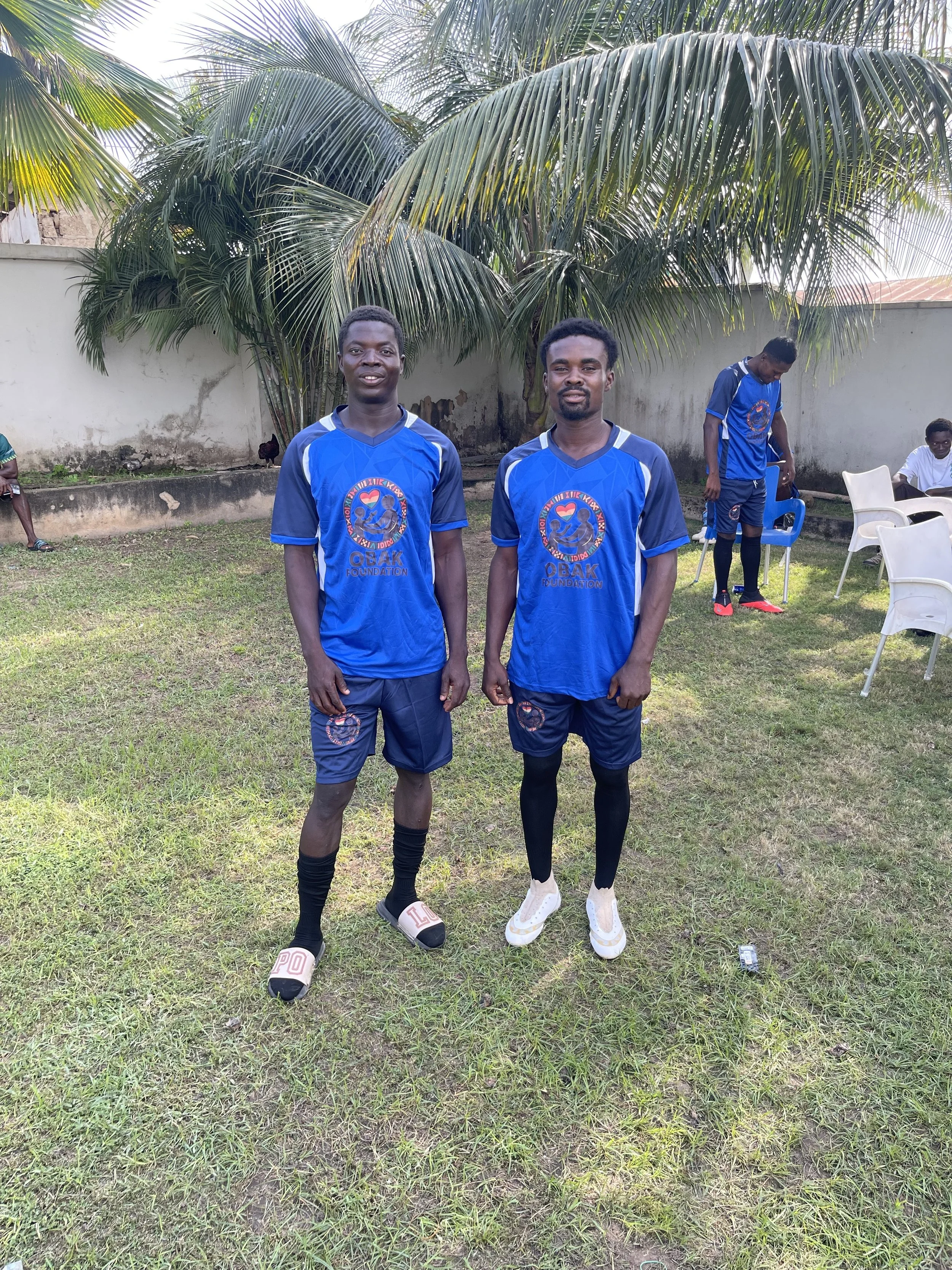Two young men standing on grass at a backyard with palm trees, wearing matching blue soccer jerseys and shorts, smiling at the camera.