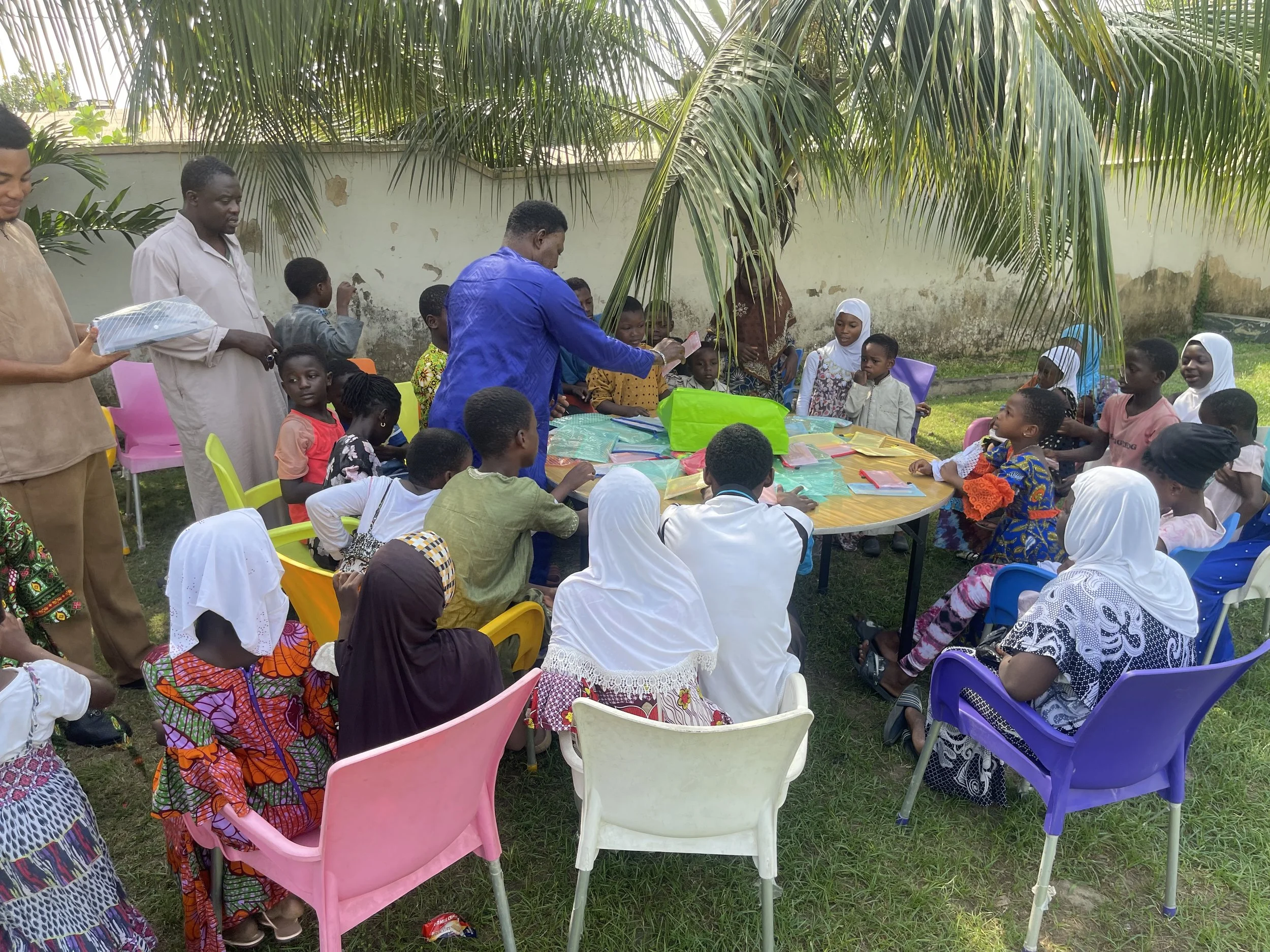 Children and adults gathered outdoors around a table under palm trees, participating in a group activity.