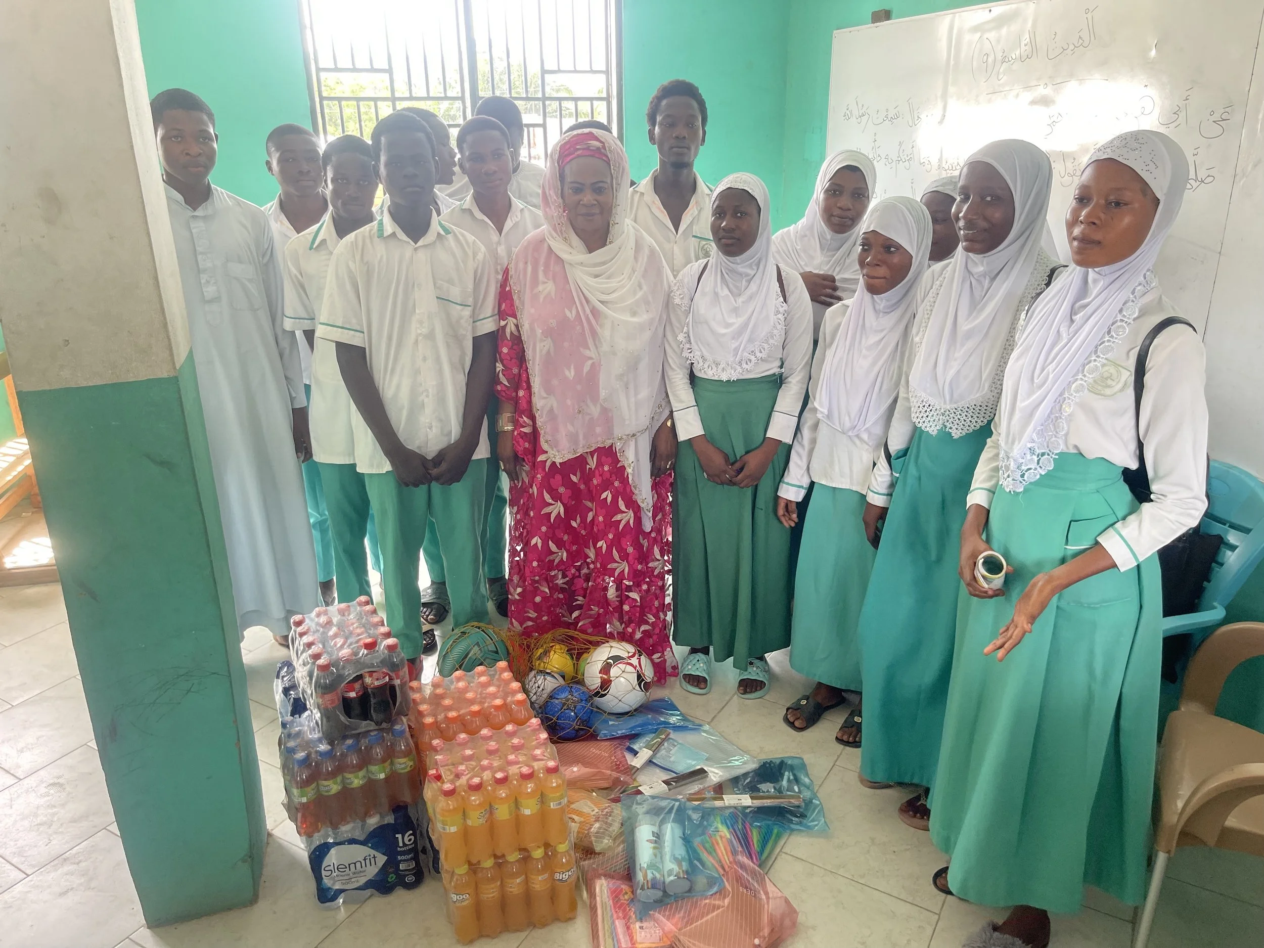 Group of students and teacher in a classroom, standing behind supplies including soda bottles, soccer balls, and school supplies.